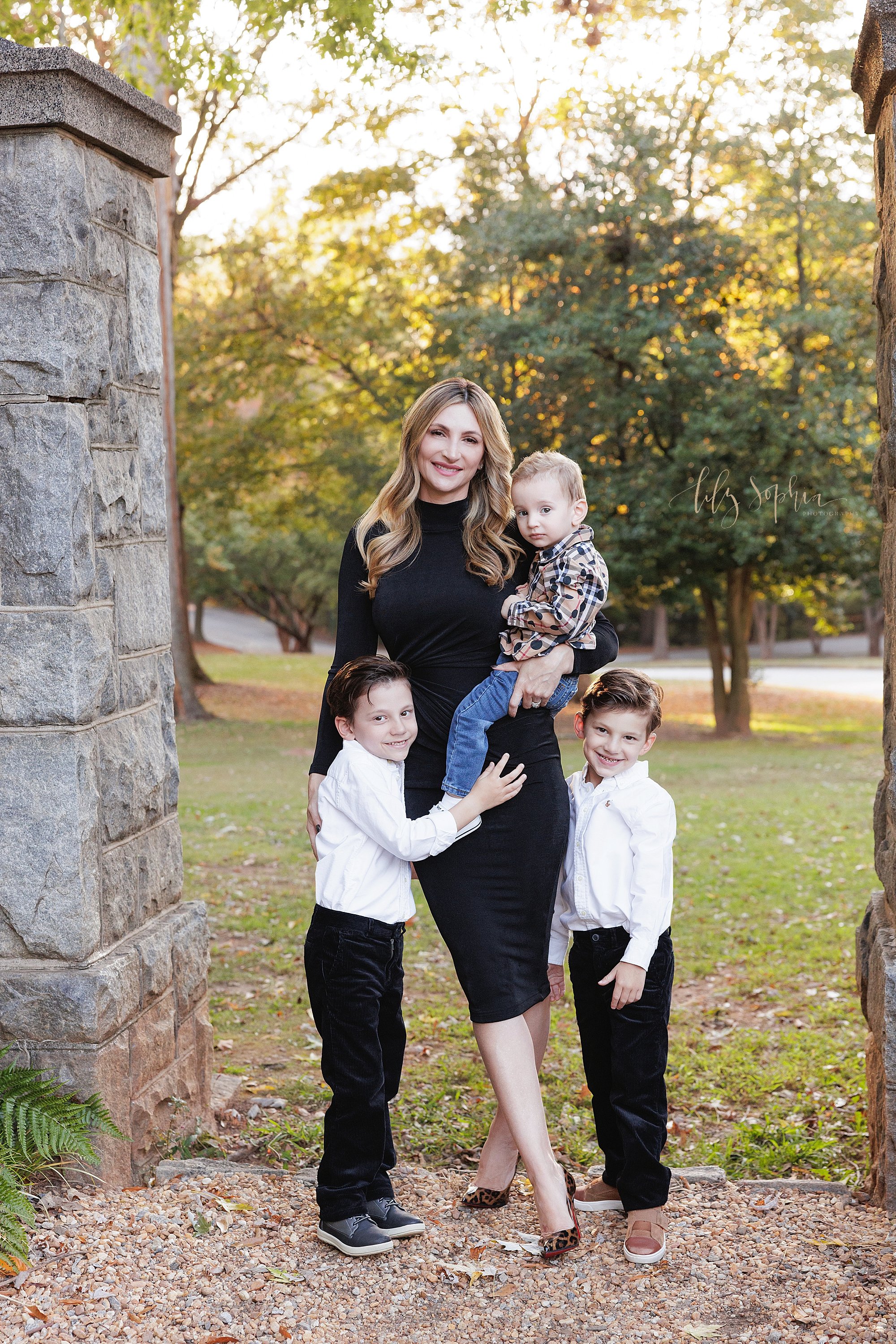  A mother stands in a stone entranceway of an Atlanta park holding her youngest son in her arms as her other two sons flank her taken for a mommy and me photo session during the fall season at sunset. 