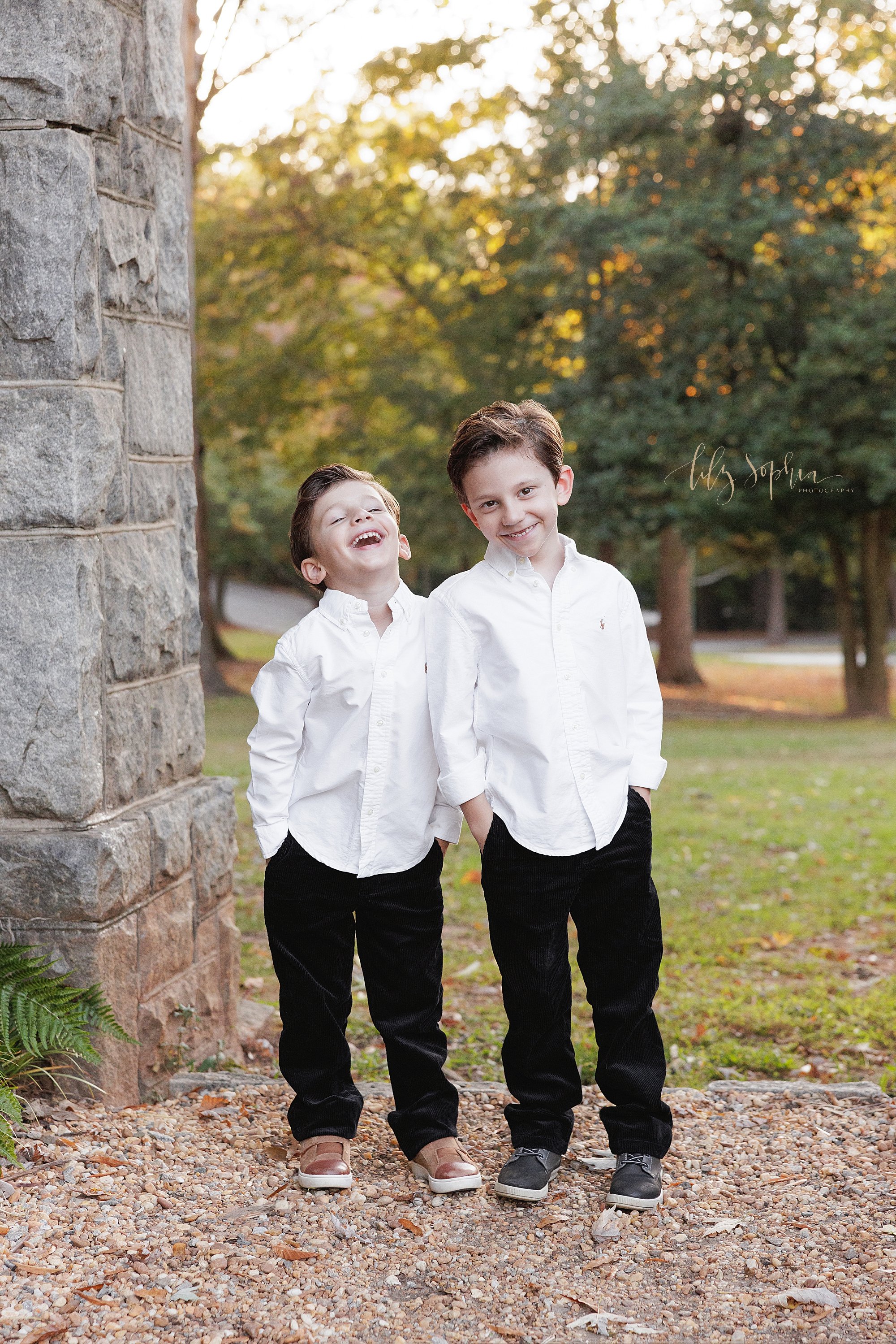  Sibling photo during a family photo shoot of two brothers standing side by side with the younger laughing taken in a stone entranceway at an Atlanta park at sunset during autumn. 