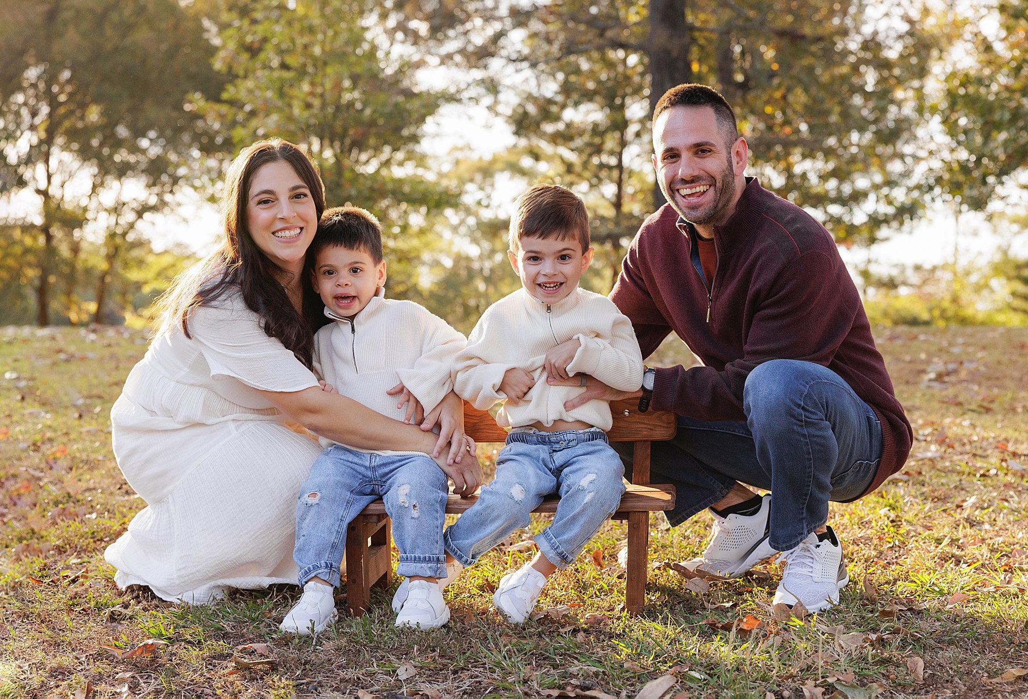  Family photo session with boy siblings sitting on a wooden bench in a park at sunset while their parents squat on either end taken near Atlanta, Georgia. 