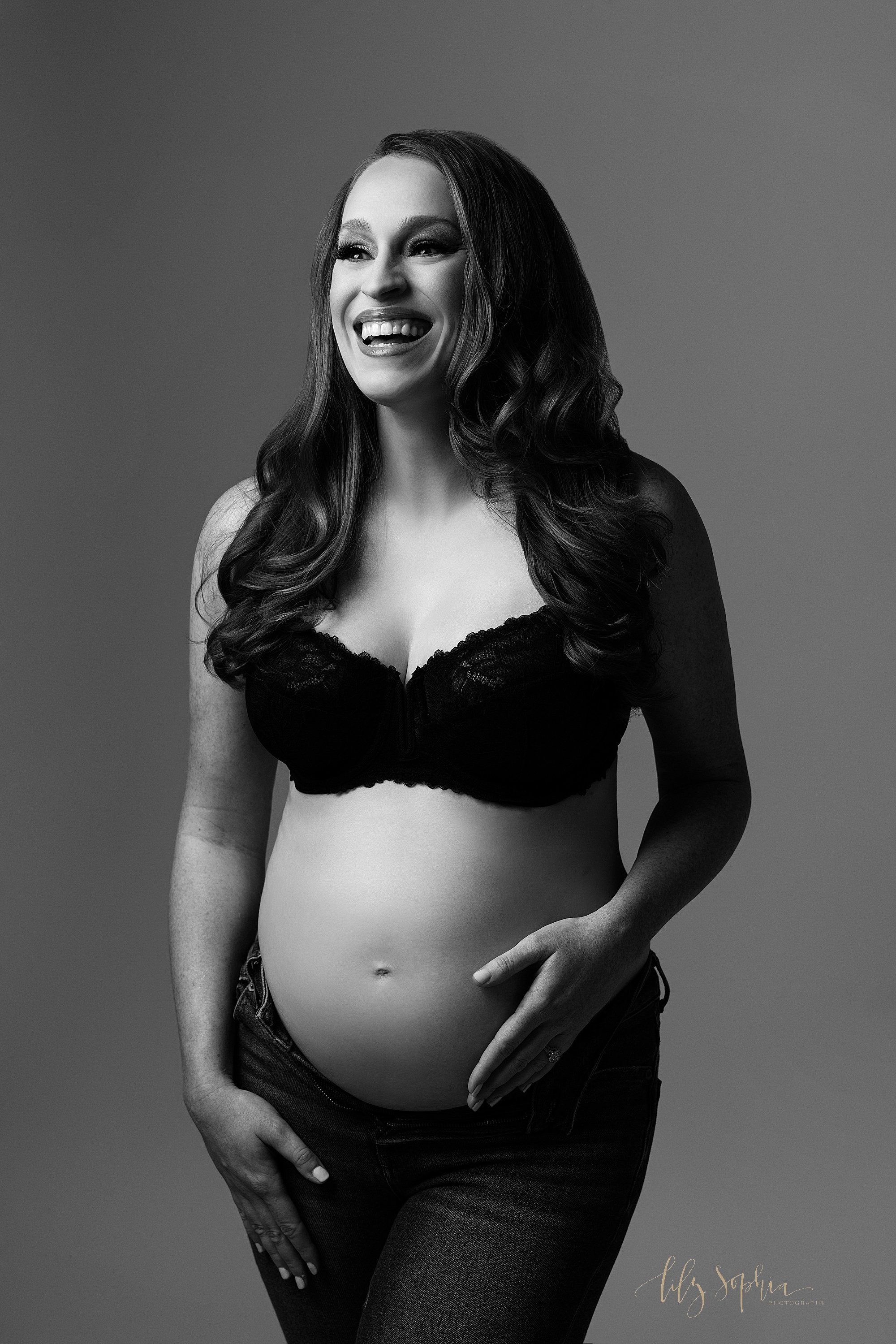  Maternity session in the modern maternity style with editorial lighting as the woman glows with a huge smile on her face as she stands in a photography studio wearing a black lace bra and unzipped jeans with her left hand on her bare belly and her r