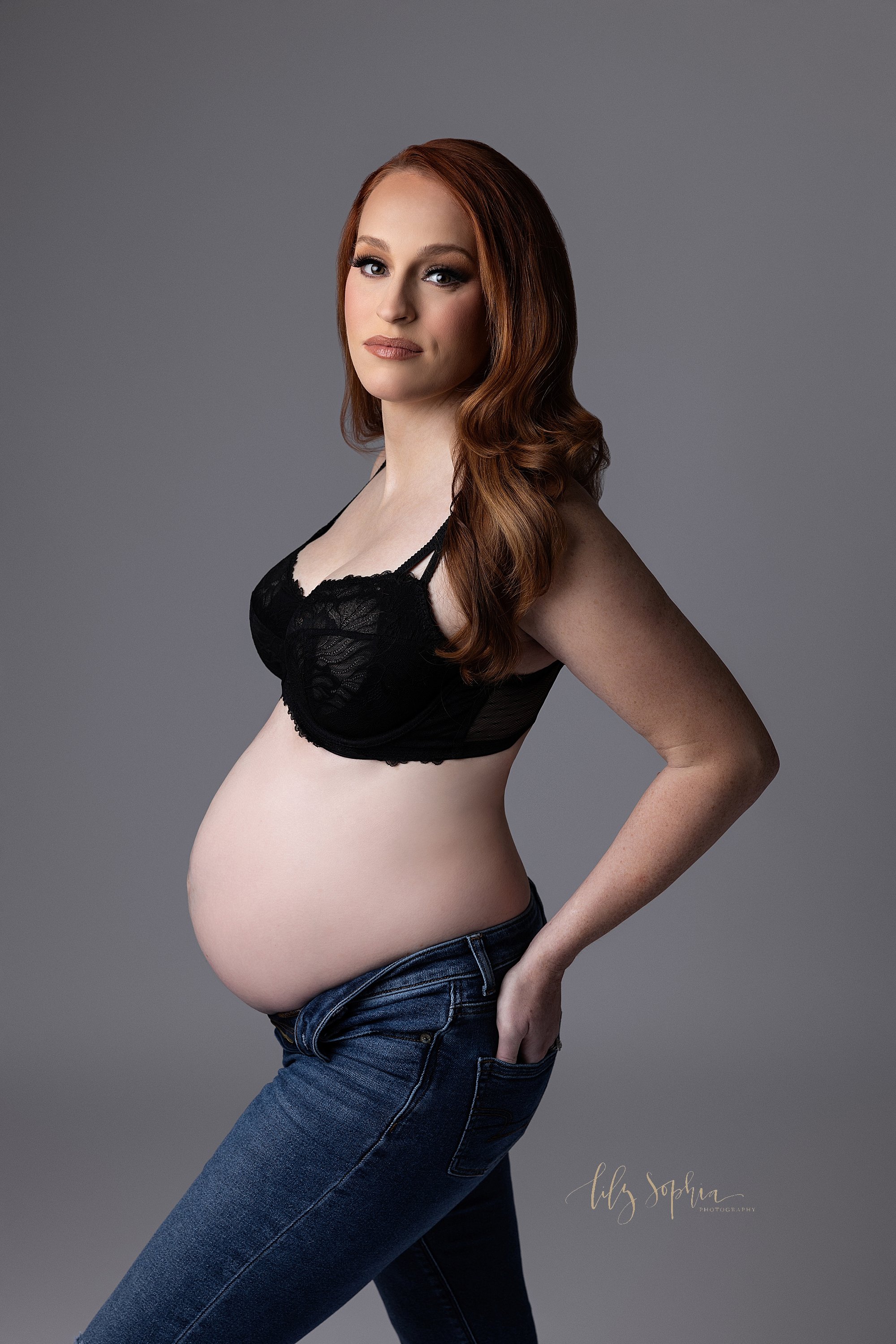  Fashion lighting is used in this maternity session using the modern maternity style as the red-headed pregnant woman stands to show the profile of her bare belly wearing a lace black bra and an unzipped pair of blue jeans with her left hand in her b