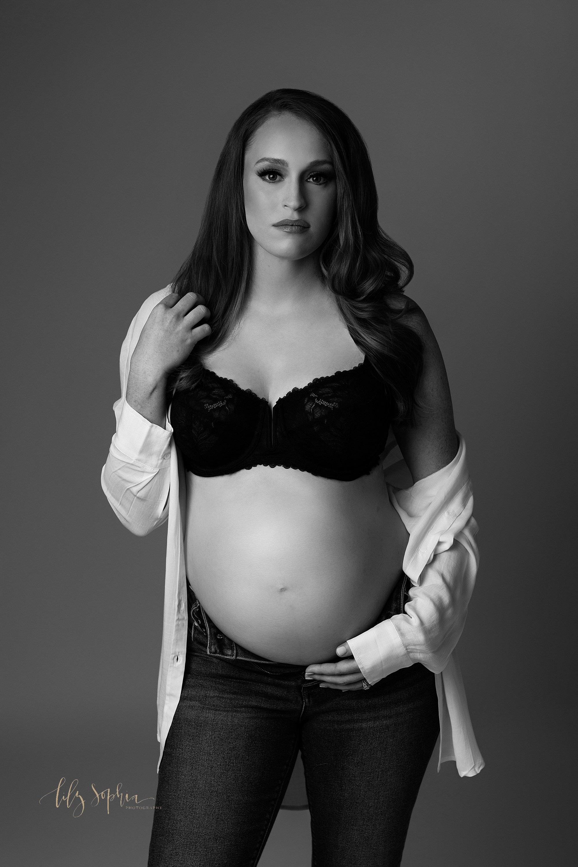  Modern maternity session in a photography studio near Old Fourth Ward in Atlanta in black and white using fashion lighting with a pregnant woman standing wearing unzipped jeans, a black bra, and a white linen shirt with the left sleeve at her elbow 