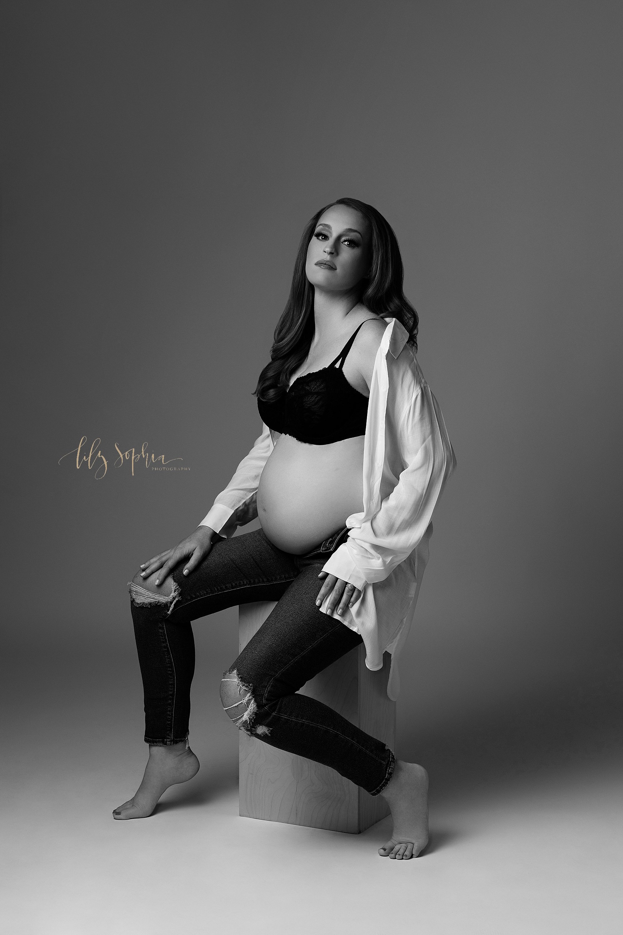  Black and white modern maternity portrait of a pregnant woman as she sits on a wooden cube wearing unzipped ripped jeans, a black bra and a white linen shirt unbuttoned and slipping off the woman’s left shoulder taken in a photography studio near Mo