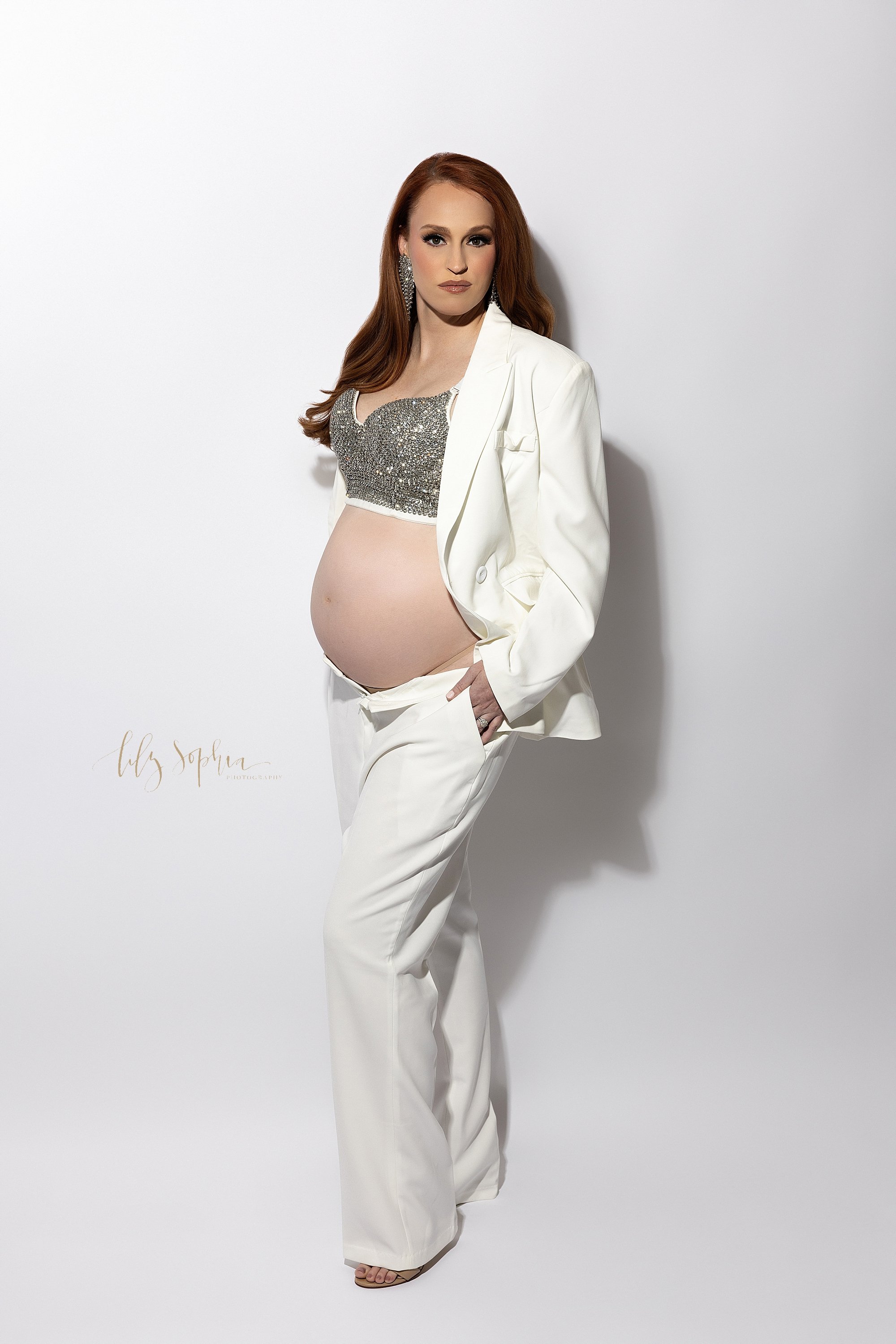  Modern maternity portrait with a red headed pregnant woman wearing a sequined bra and a white suit jacket with  white opened pants to show her bared belly as she places her hands in her pants pockets taken in a photography studio near Oakhurst in At