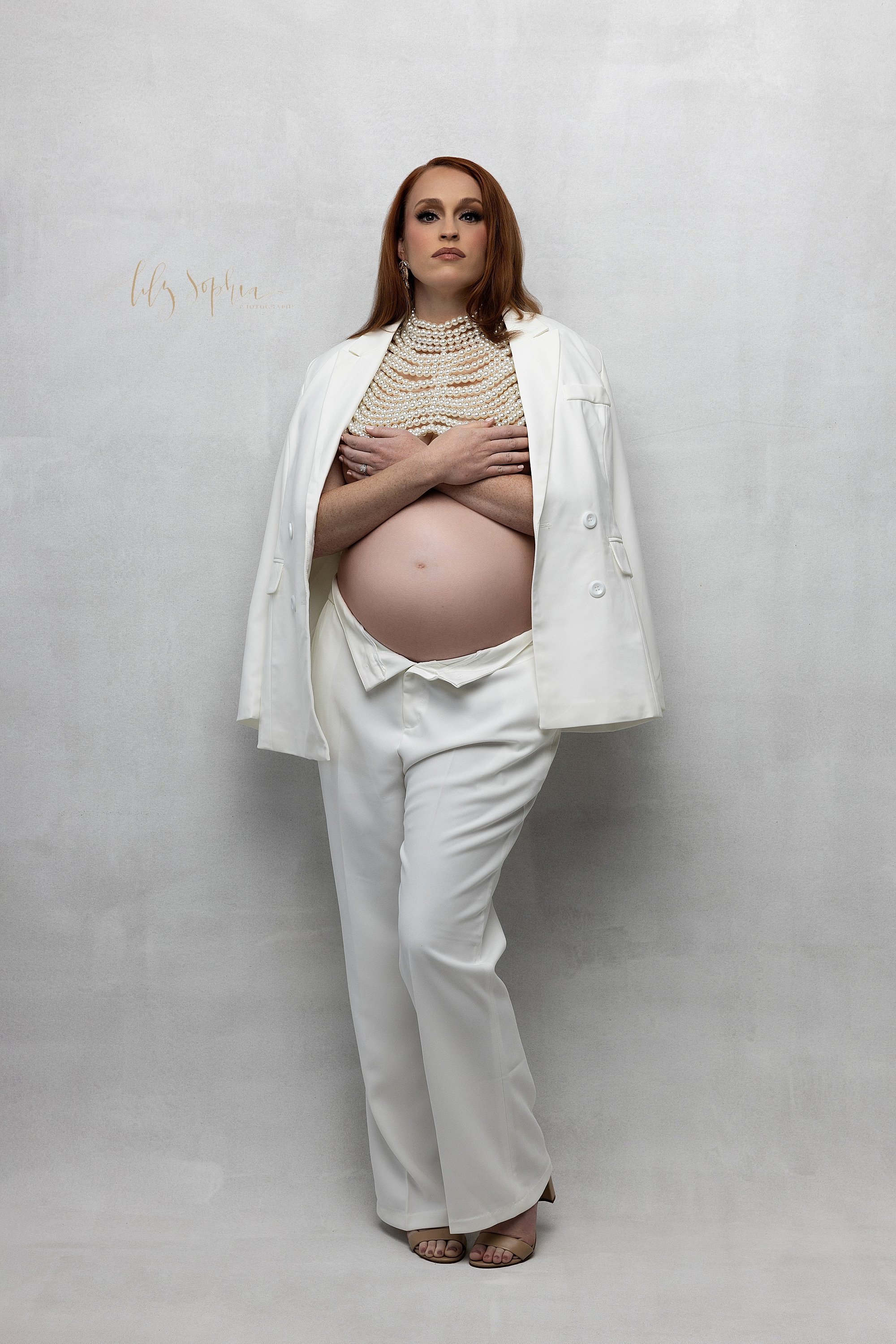 Modern maternity photo session with a pregnant woman wearing unbuttoned white pants a pearl collar and a suit jacket draped over her shoulders as she places her hands over her bare breasts and bares her belly in a photography studio near Kirkwood in