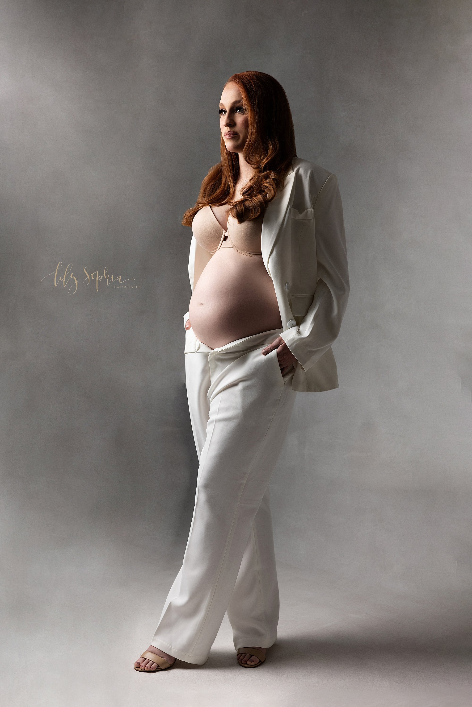  Modern maternity photo shoot using studio lighting with a red haired mother as she stands in a white suit with the jacket open to reveal her bra and her bare belly with the pants unzipped taken in a photography studio in Ponce City Market in Atlanta