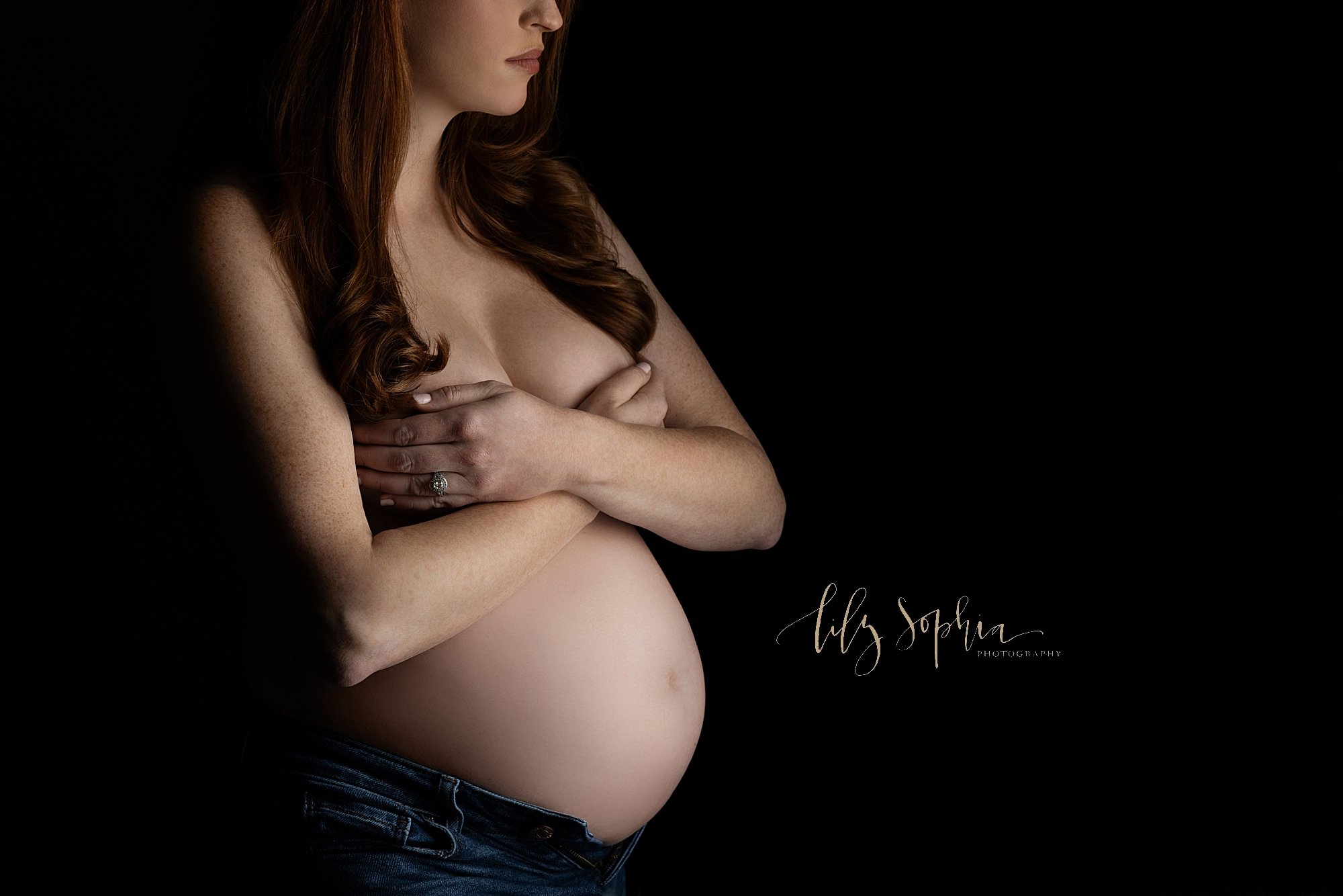  Maternity portrait of a red haired mother as she holds her breasts and bares her belly while wearing unzipped blue jeans taken in Midtown, Atlanta in a natural light photography studio. 