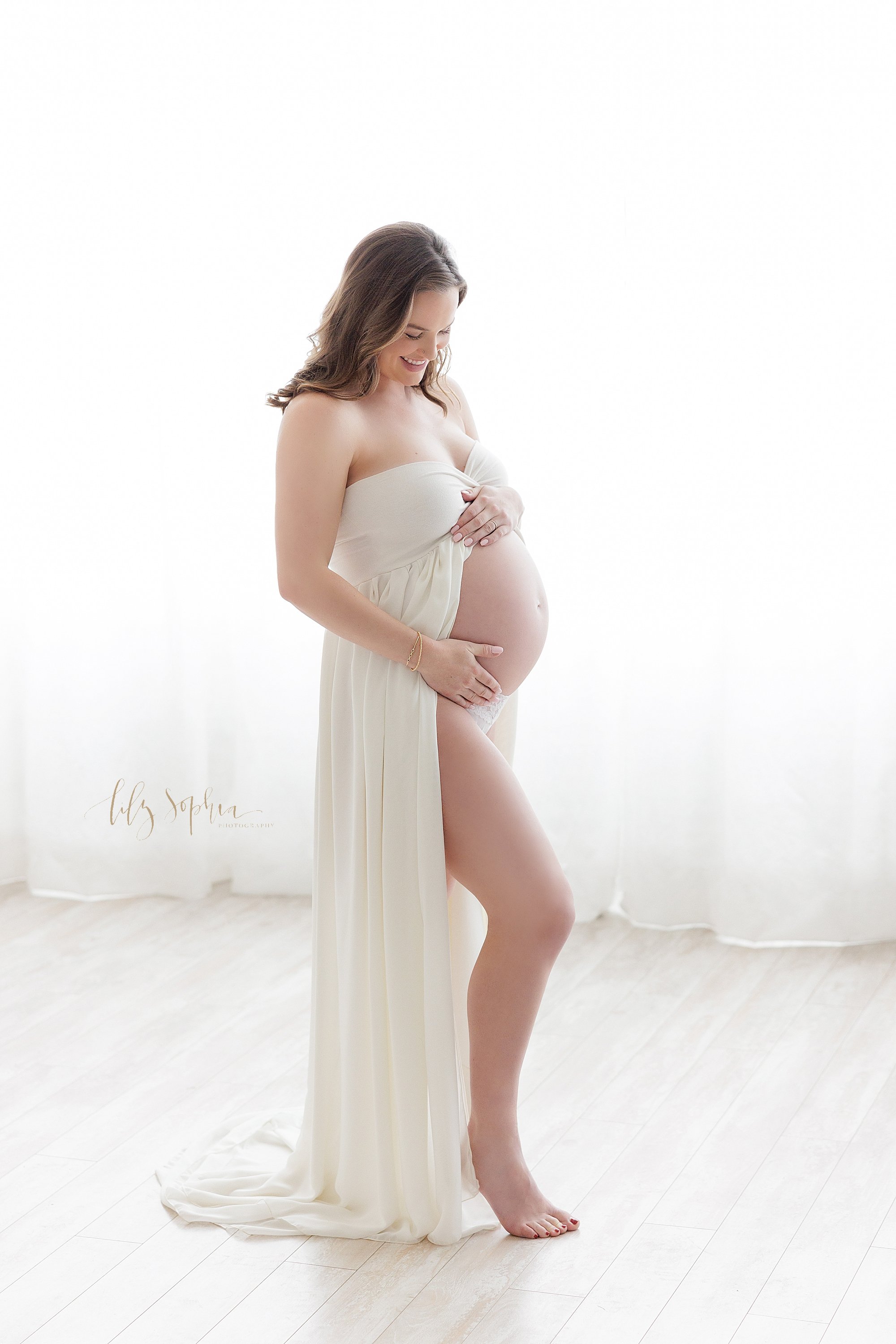  Maternity portrait taken in front of window streaming natural light of an expectant mother as she stands in a split front bandeau gown with her right leg bent in front of her left as she frames her belly while smiling as she thinks about her upcomin