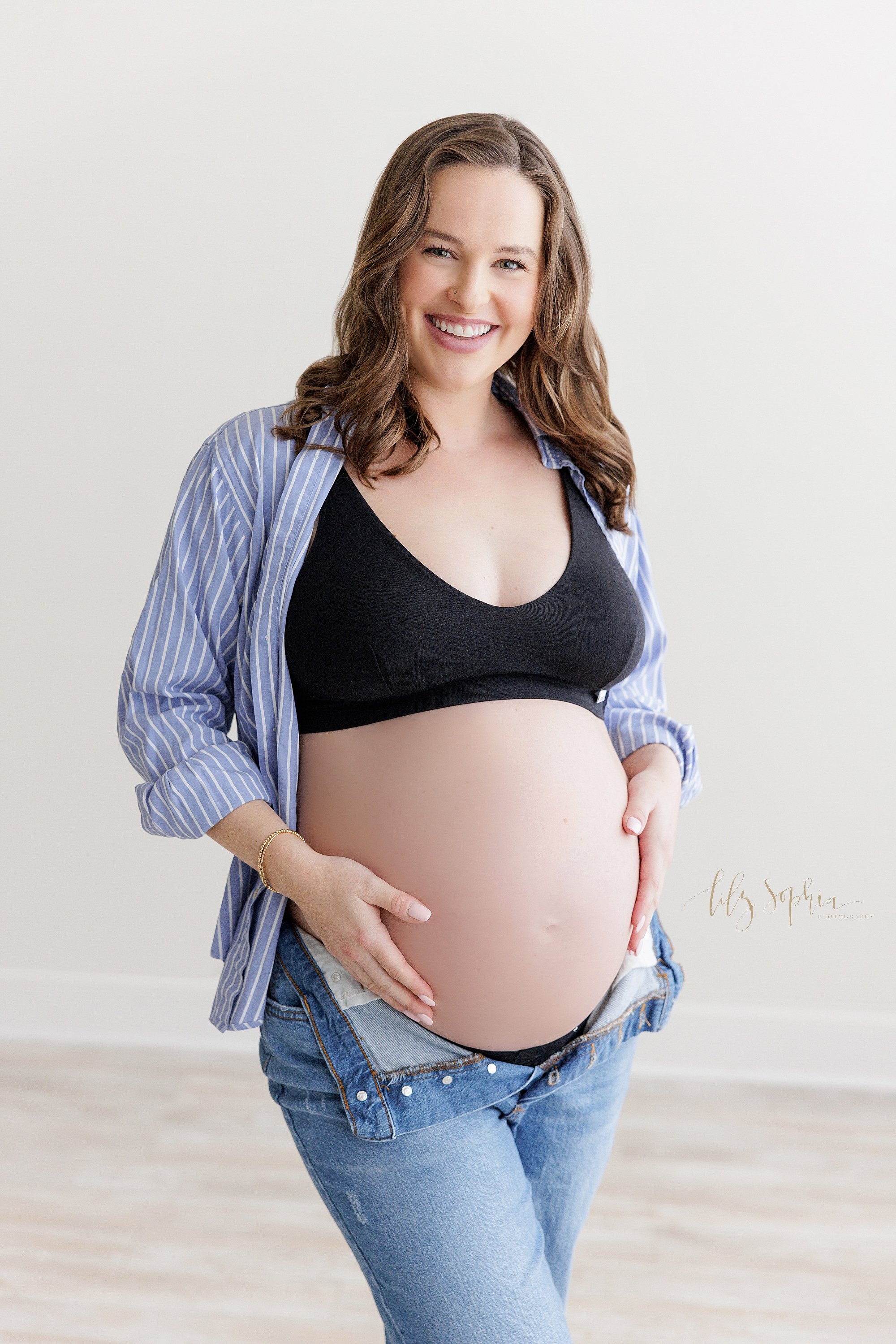  Casual maternity photo session with a young mother as she stands in a studio wearing a pair of blue jeans, a black bra, and an opened button down blue and white striped chambray shirt with her bare belly showing as the blue jeans are unbuttoned and 