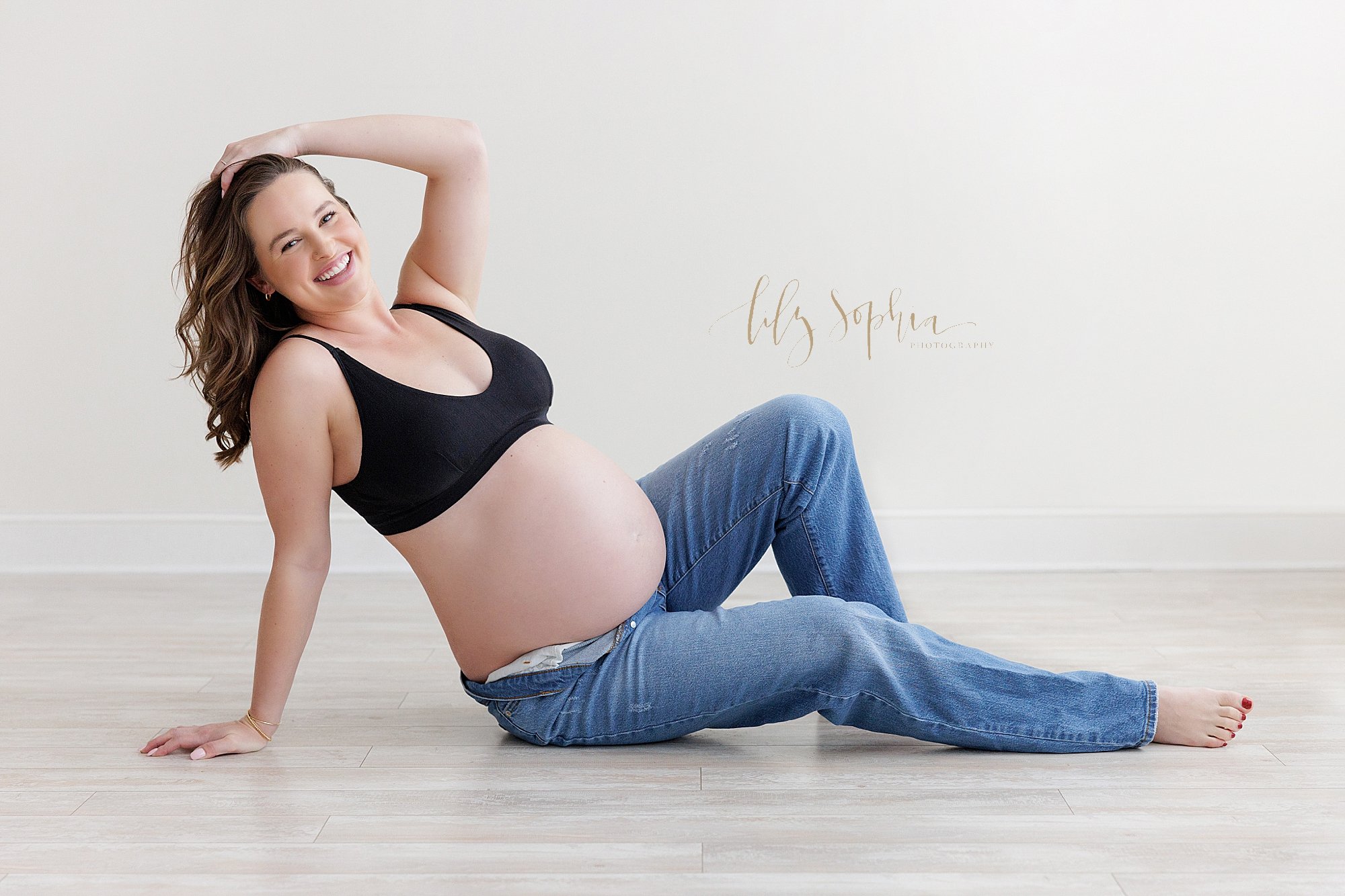  Maternity photo session in a natural light studio with an expectant mother as she sits on the floor of the studio wearing a black bra and blue jeans unbuttoned to bare her belly as she leans with her right hand on the floor and her left hand on the 