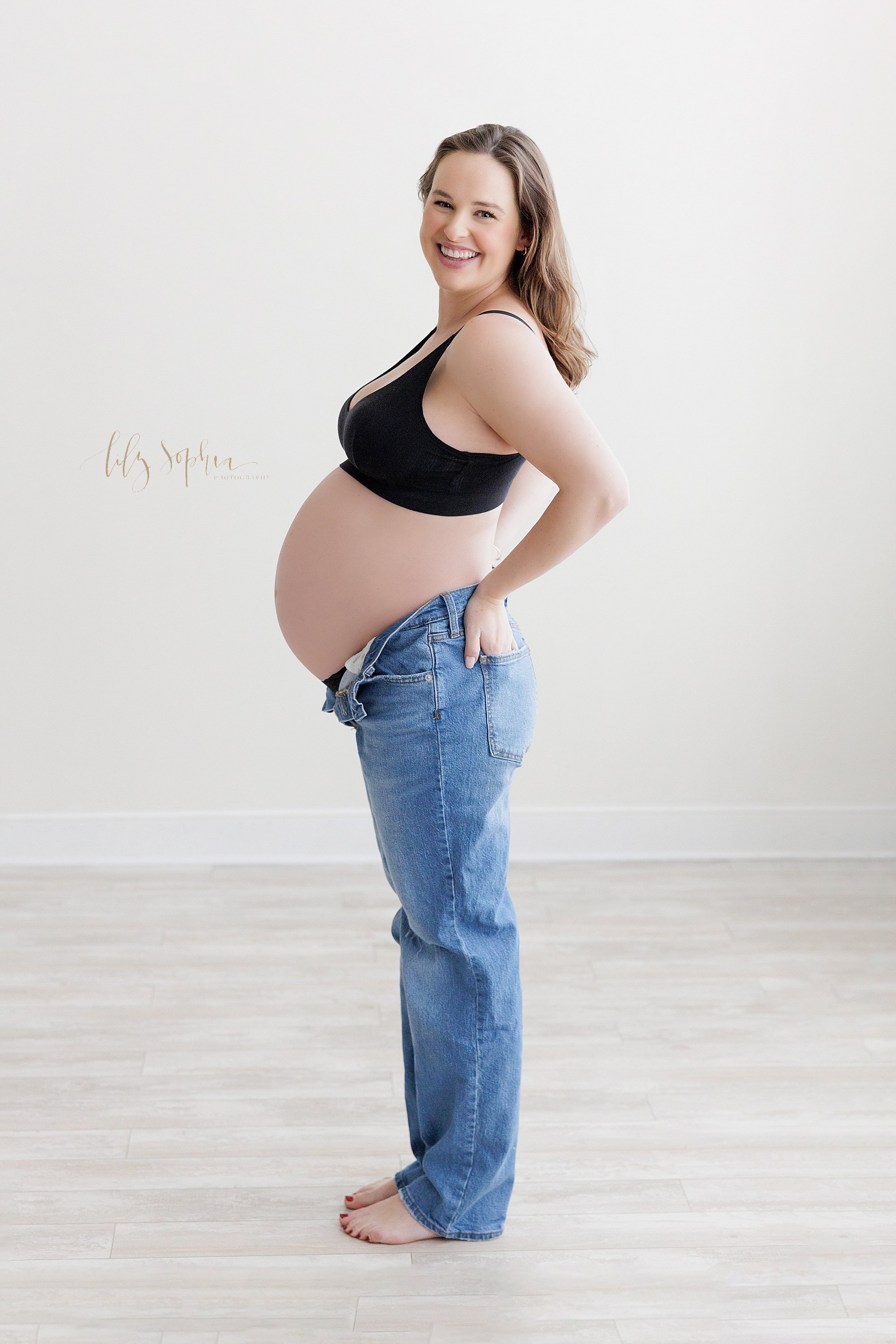  Maternity portrait of an expectant mother wearing a  black bra and a pair of blue jeans that are unbuttoned to show her bare belly as she looks over her left shoulder while putting her hands in the back pockets of her jeans as she stands in a natura