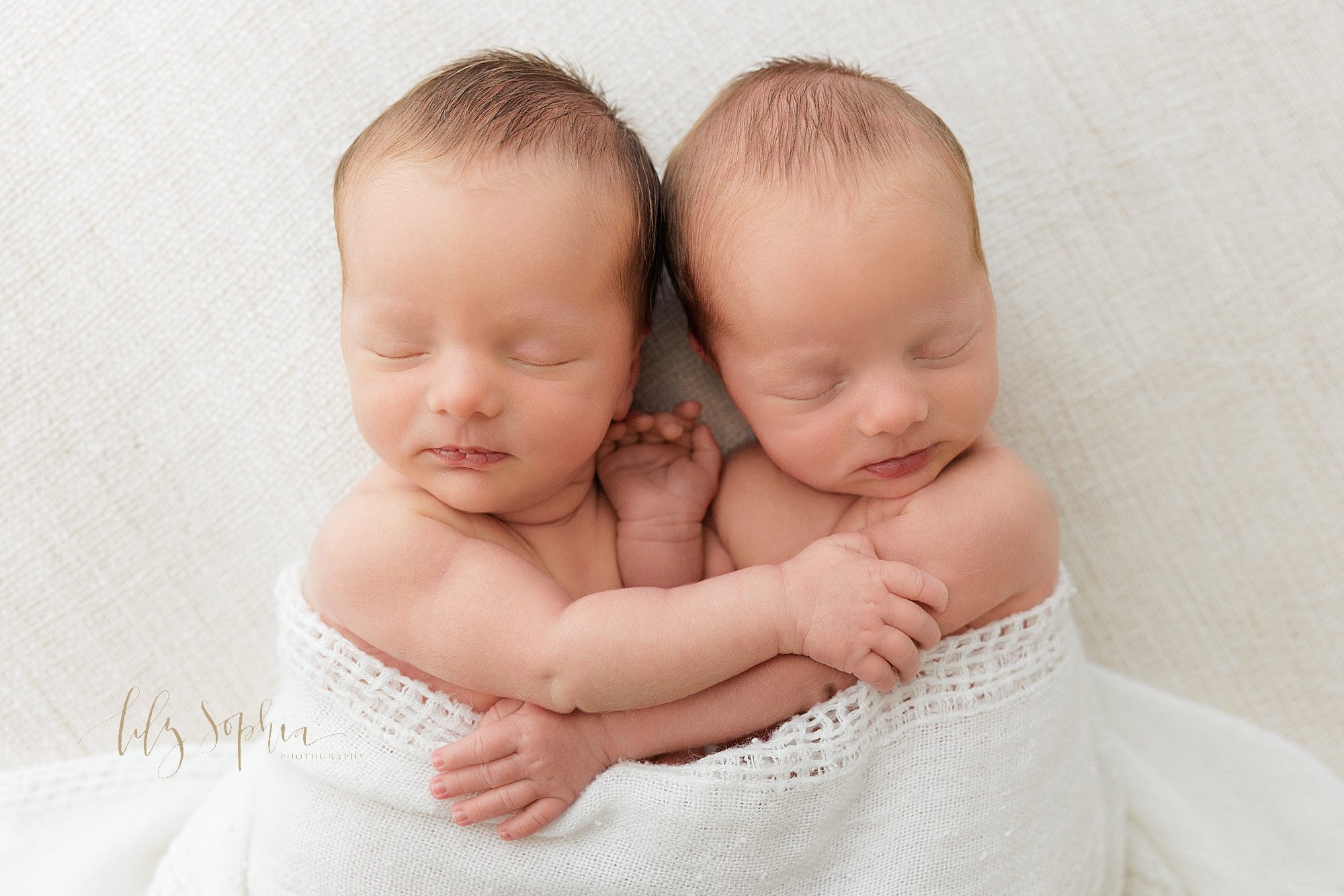  Identical twin newborn baby boys lie next to one another wrapped in a single soft blanket as one of the boys grabs his brother’s arm to hug him taken using natural light near Smyrna in Atlanta in a photography studio. 