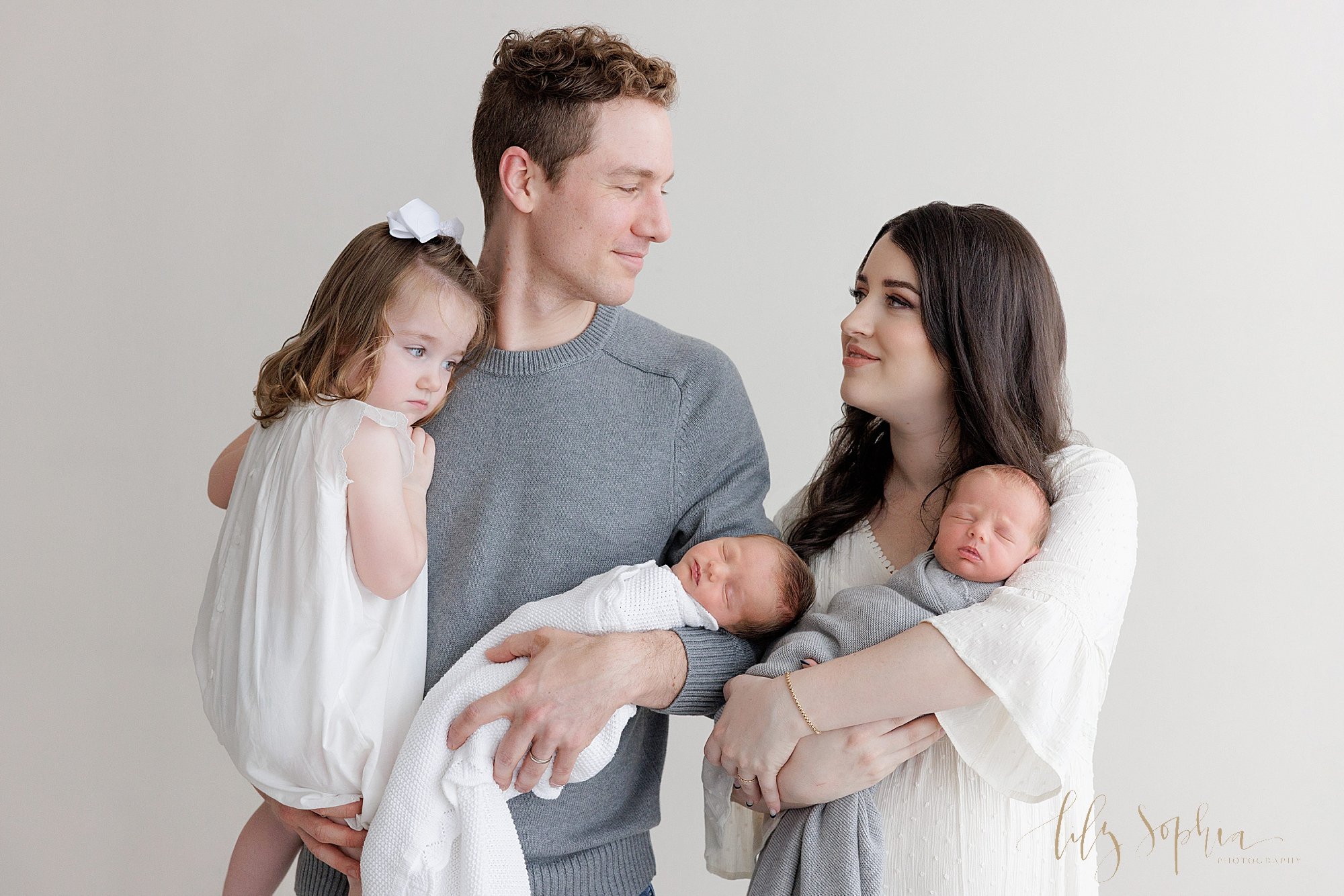  Family newborn portrait of a father holding his young daughter in his right arm and one of his newborn identical twin sons in his right arm as mom stands to his right and holds their other son in her left arm while the couple look lovingly at one an