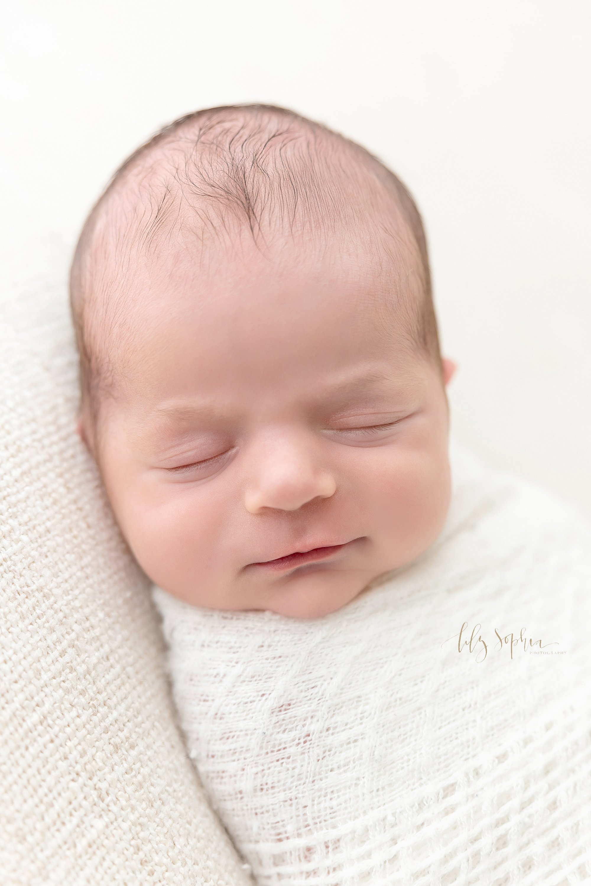  Portrait of a newborn baby boy as he peacefully sleeps and smiles in his sleep while being swaddled to his chin in a soft white blanket taken in a natural light studio near Ansley Park in Atlanta, Georgia. 