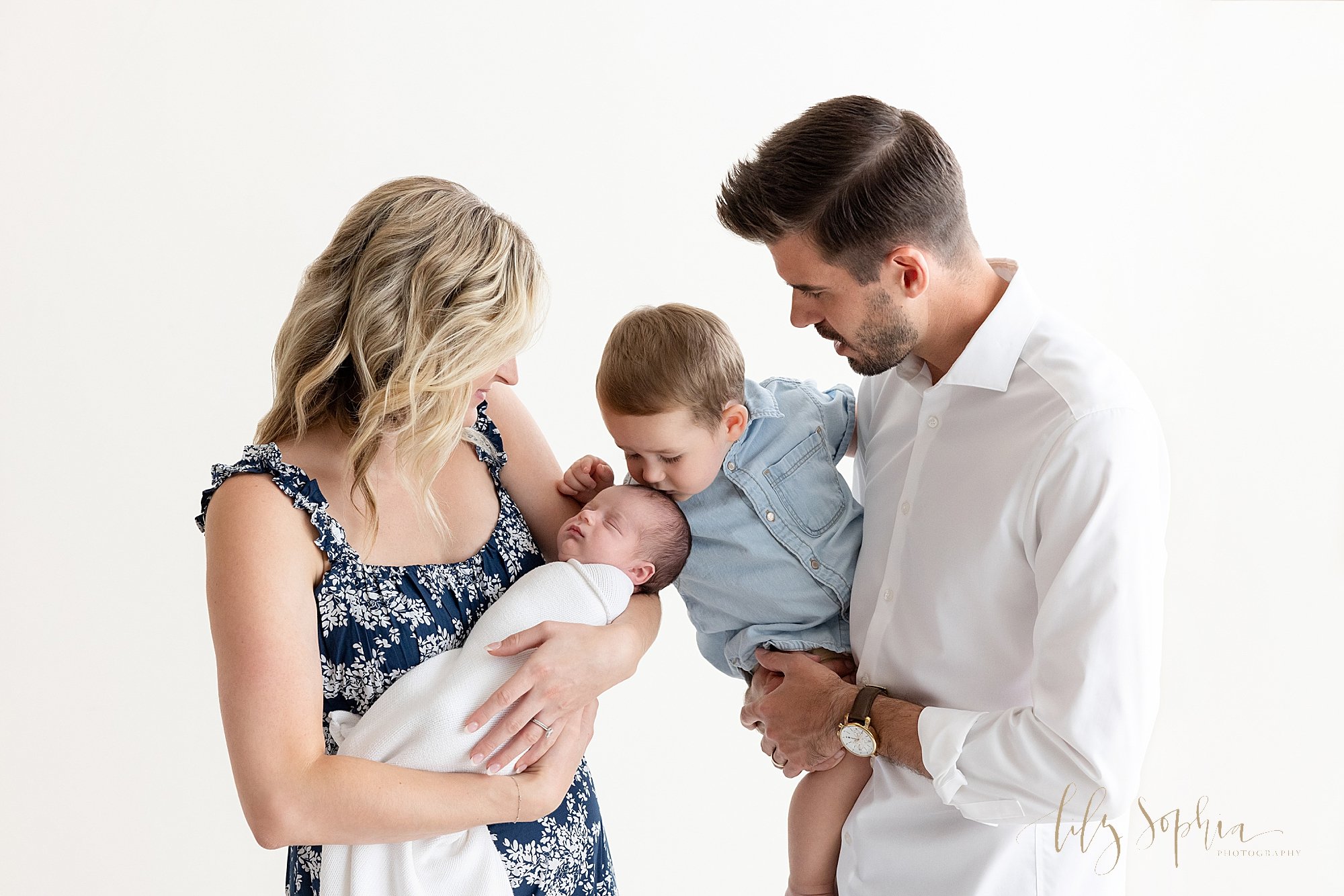  Family newborn photo session with mom cradling their newborn son in her arms as dad faces mom holding their young son who kisses the crown of his brother’s head taken next to a window streaming natural light in a studio near Vinings in Atlanta. 