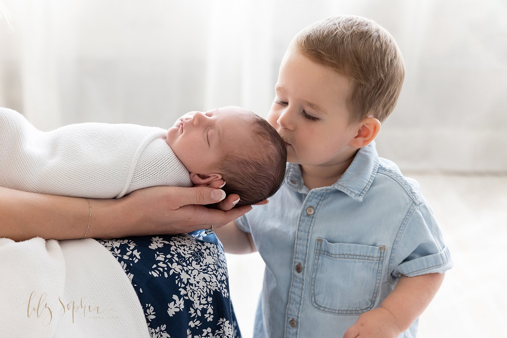  Precious newborn portrait of a sleeping newborn baby boy being held on his mother’s lap with his head in her hands as his toddler brother plants a kiss on the crown of his head taken in front of a window streaming natural light in a photography stud