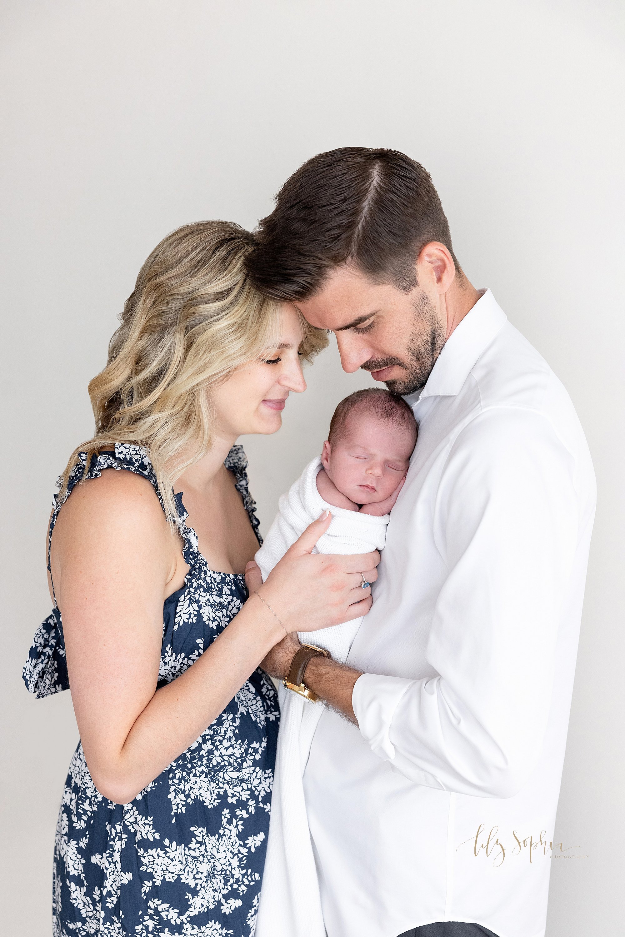  Newborn family portrait of dad holding his newborn son against his chest as he sleeps and mom takes her hands to hold her son as she faces her husband as they stand in a photography studio near Morningside in Atlanta that uses natural light. 