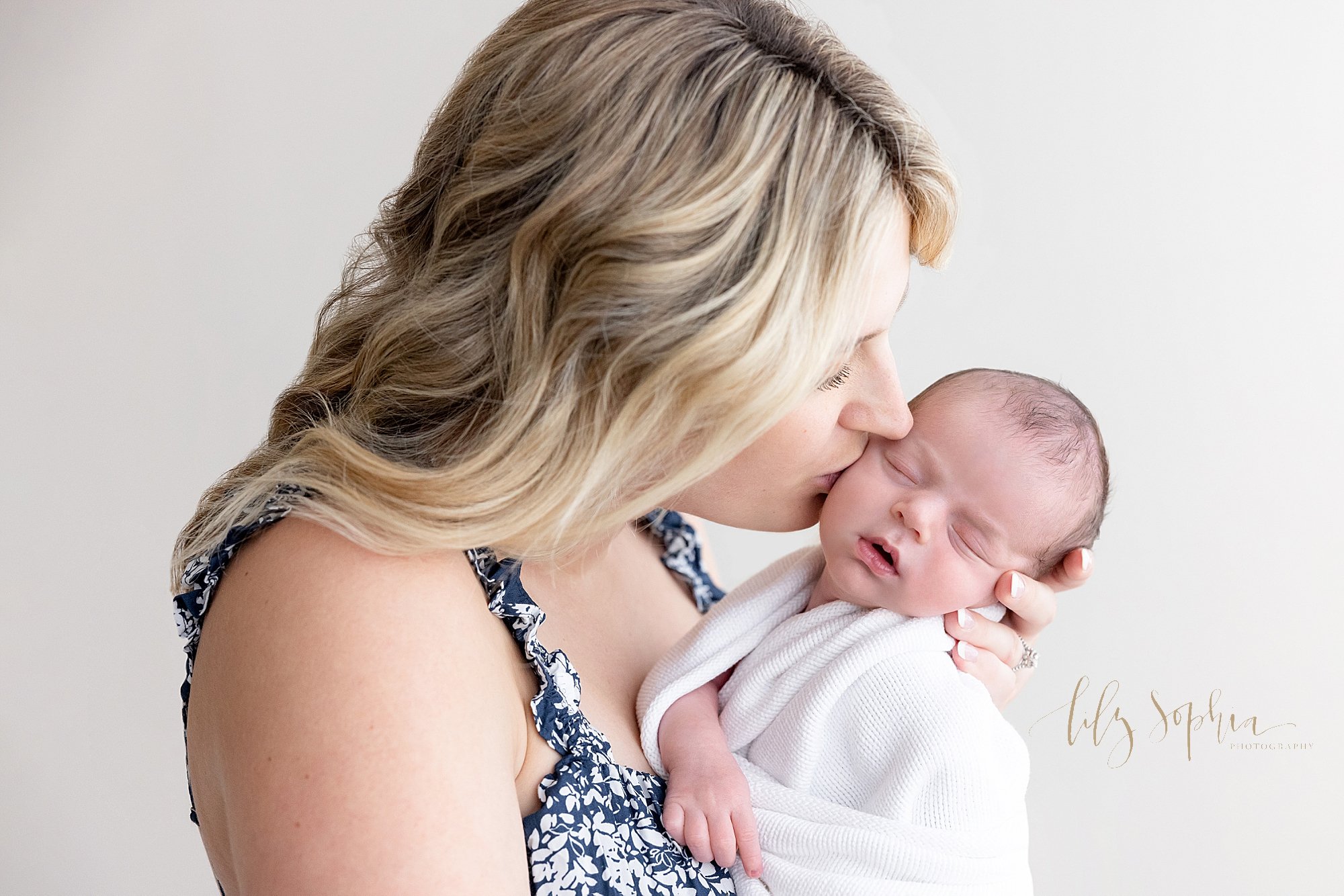  Sweet moment for a newborn and his mother as she holds him with his right hand sticking out of blanket as mom holds his head in her hand and kisses him on the cheek while she stands in a natural light studio near Kirkwood in Atlanta. 