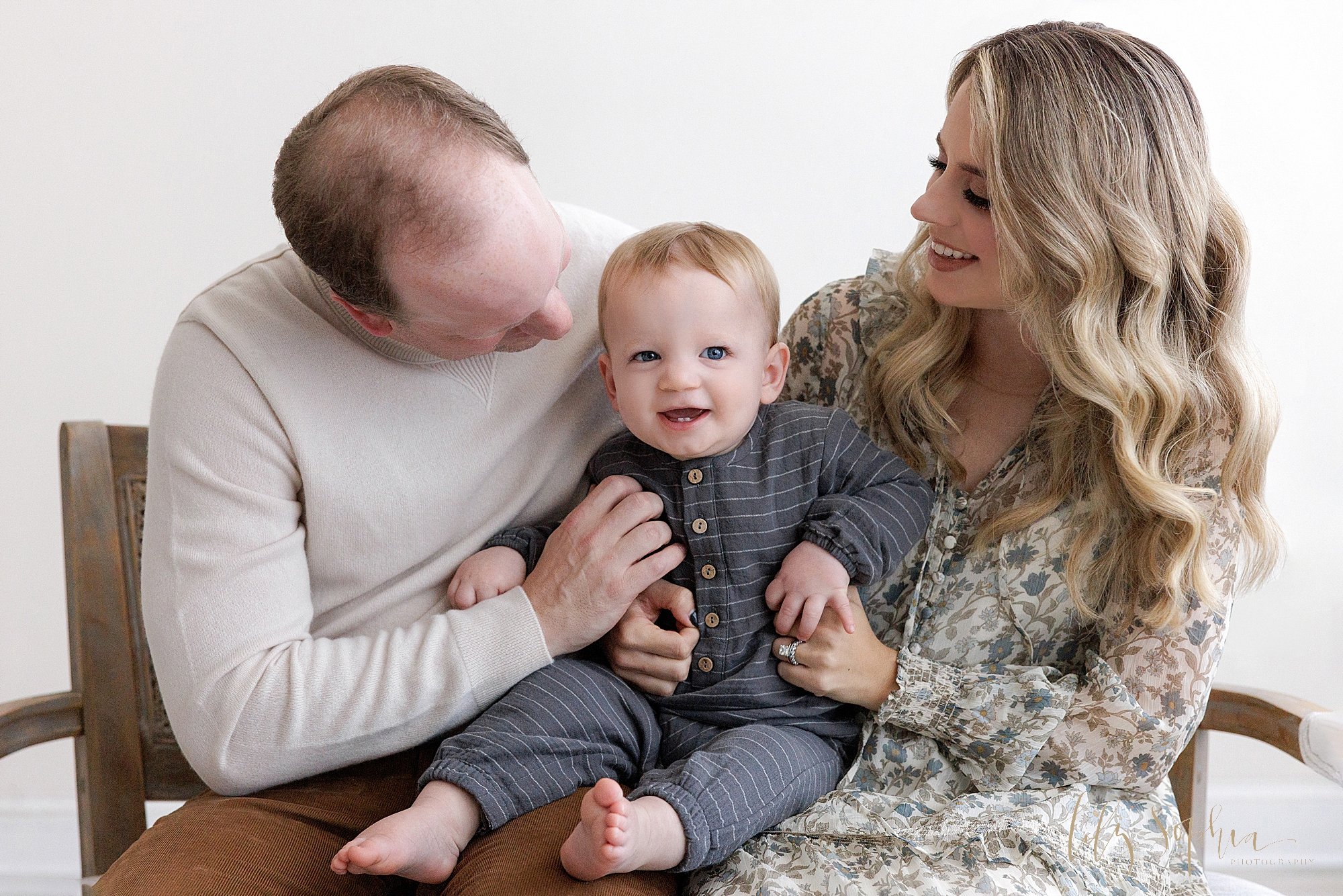  Family first birthday photo session as mom and dad sit next to one another on a bench with their one year old son on their laps as dad tickles him to make him laugh and the parents look toward their son taken near Buckhead in Atlanta in a photograph