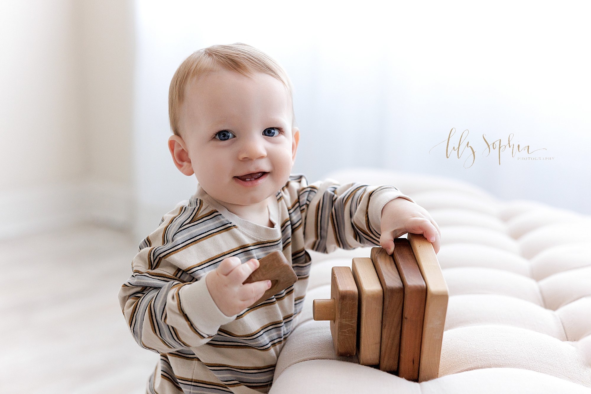  First birthday portrait of a one year old baby boy as he stands next to a tufted bench while playing with a wooden stacking toy as he turns his head to the right taken in front of a window streaming natural light near Kirkwood in Atlanta in a photog