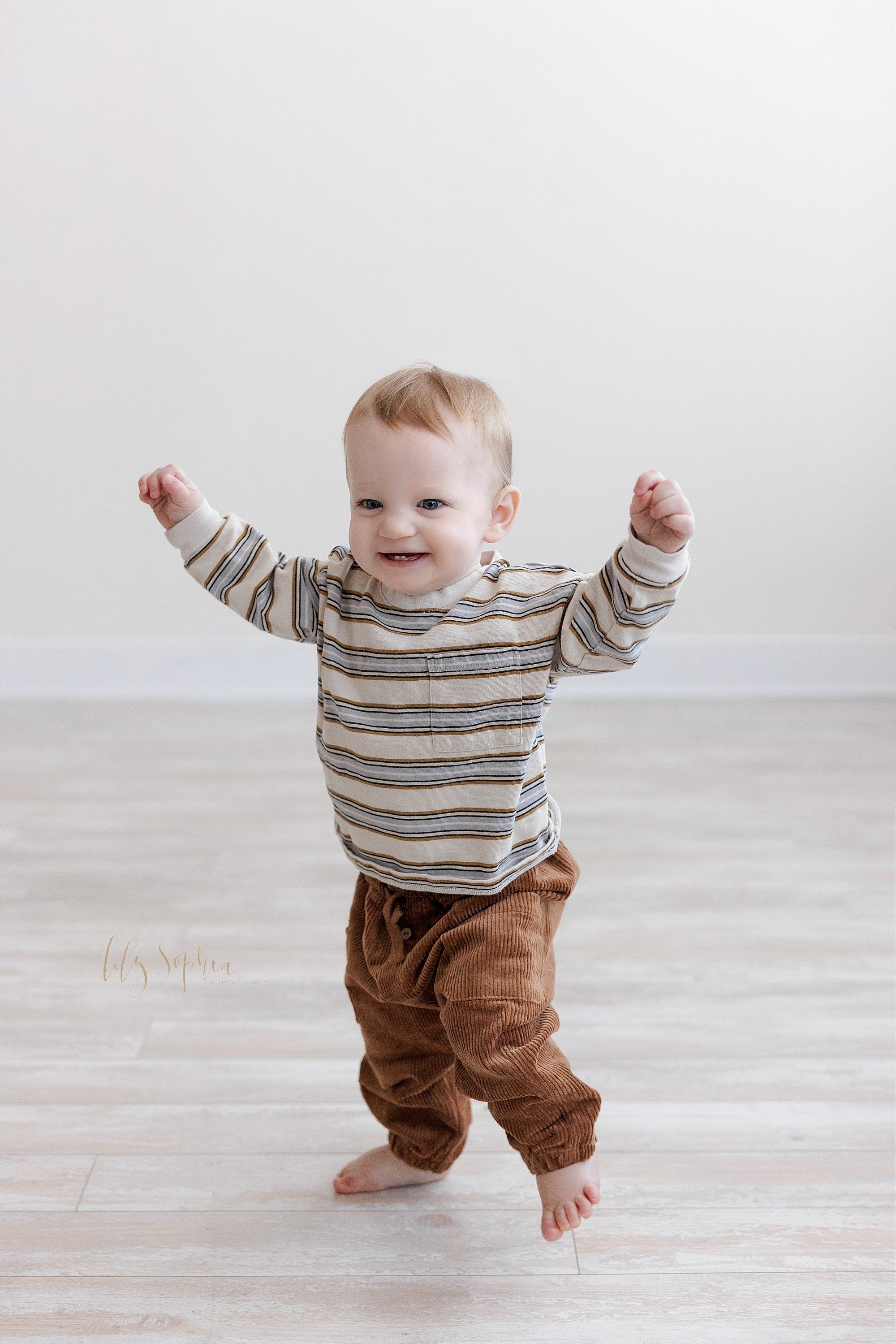  First birthday photo of an excited one year old baby boy as he raises his hands in the air, smiles to show his tiny bottom teeth, and runs barefoot in a photography studio near Old Fourth Ward in Atlanta that uses natural light. 