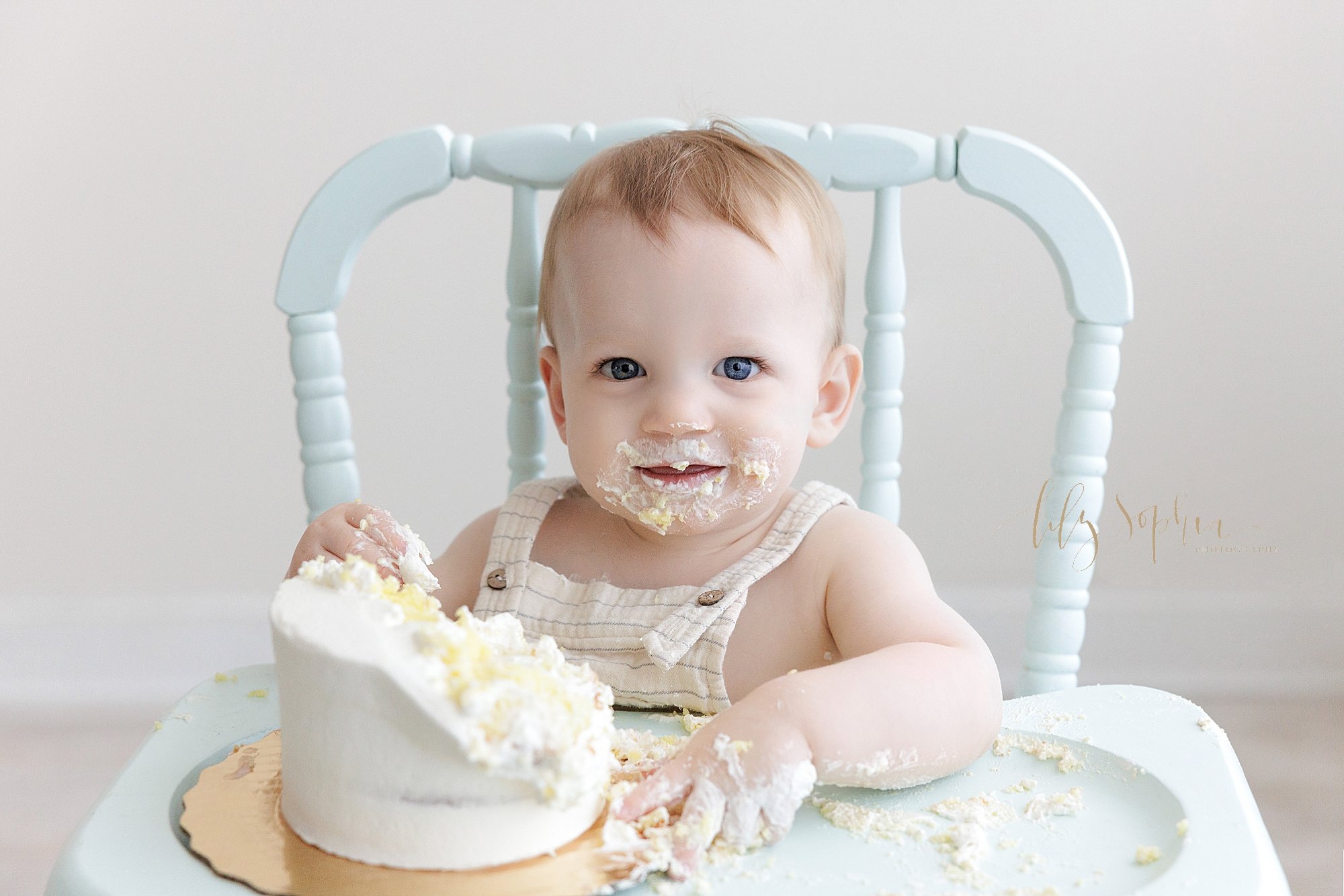  First birthday smash cake photo of a one year old boy sitting in an antique highchair with his half devoured smash cake on the tray while his hands and face are covered in icing and cake taken in a natural light studio near Poncey Highlands in Atlan