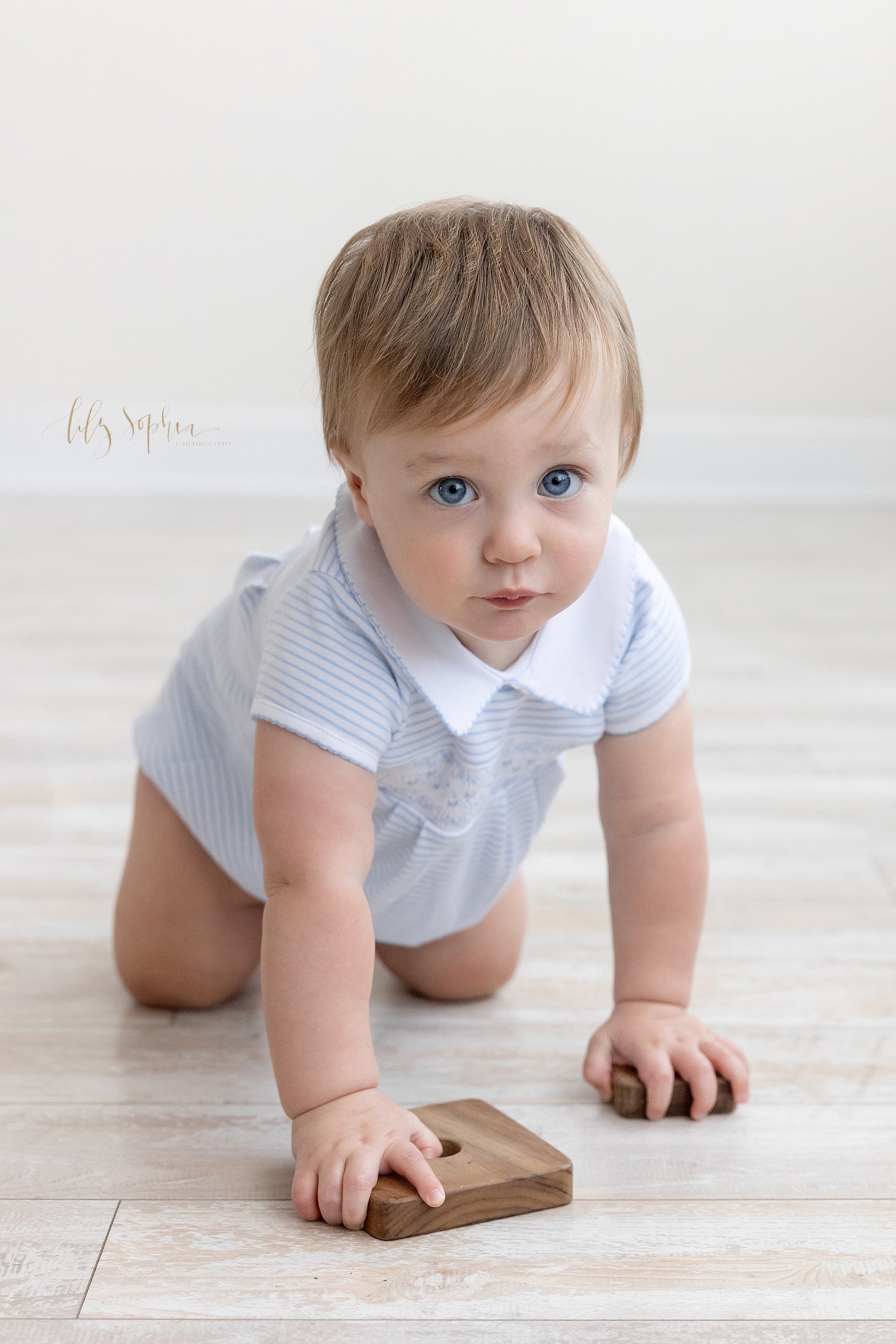  A one year old little boy crawls while holding two wooden stacking blocks in his hand along a wooden floor in a photography studio near Poncey Highlands in Atlanta that uses natural light for his first birthday photo shoot. 