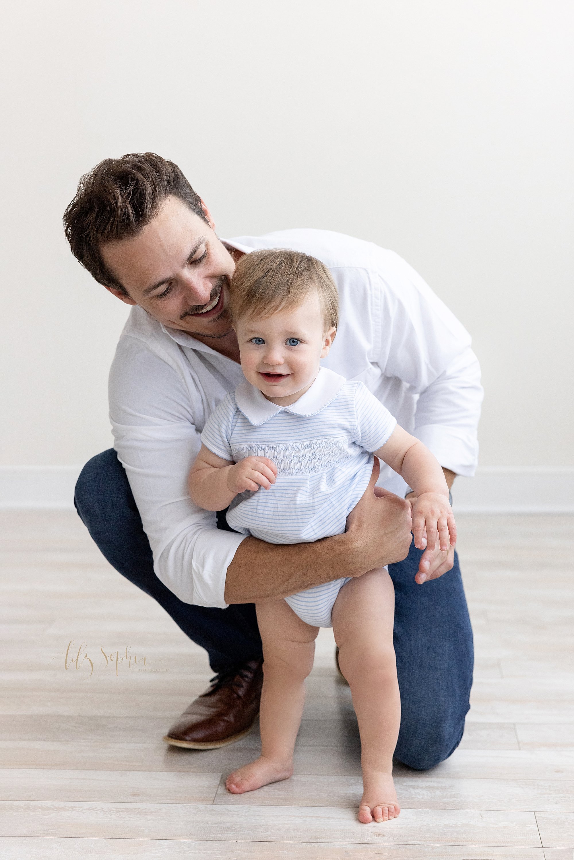  First birthday photo session with a one year old little boy as his father squats behind him with his right arm around his son’s waist to help him stand taken near Alpharetta in Atlanta in a photography studio that uses natural light. 