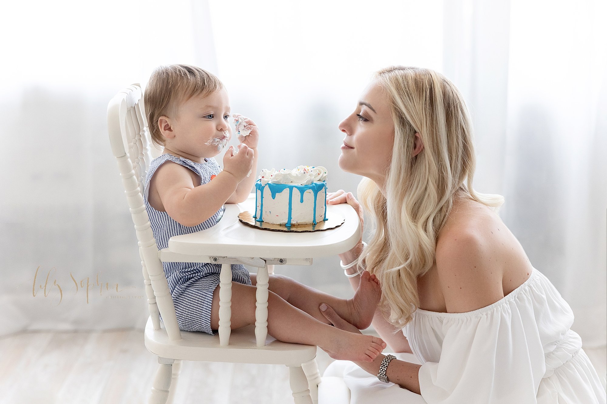 Mom squats in front of her one year old son as he sits in an antique high chair wearing icing from his smash cake on both his hands and his face as natural light streams through the window behind them in a photography studio near Decatur in Atlanta 