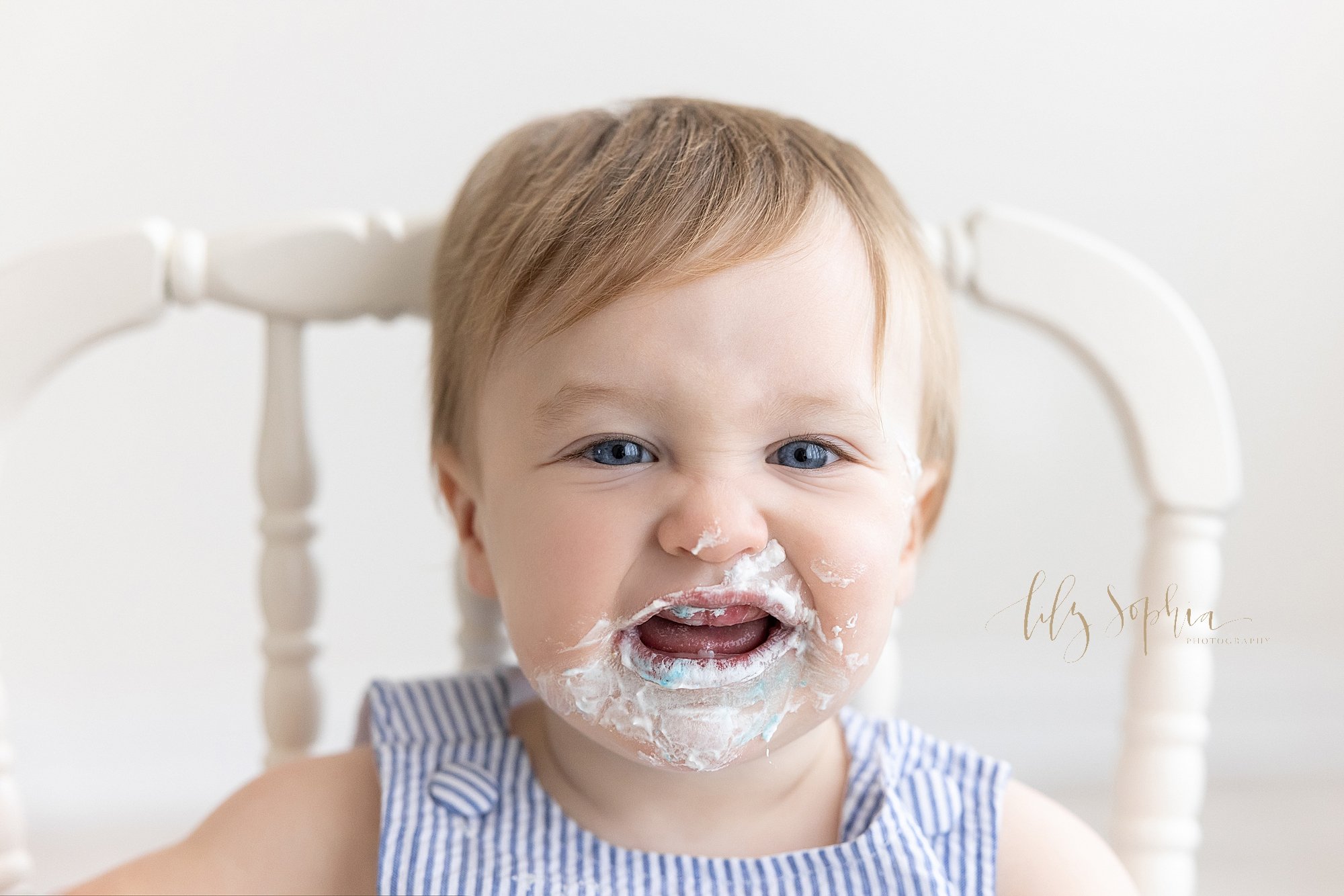  Smash cake photo session with a one year old little boy celebrating his first birthday as he sits in an antique high chair with icing surrounding his toothless grin taken near Virginia Highlands in Atlanta in a natural light photography studio. 