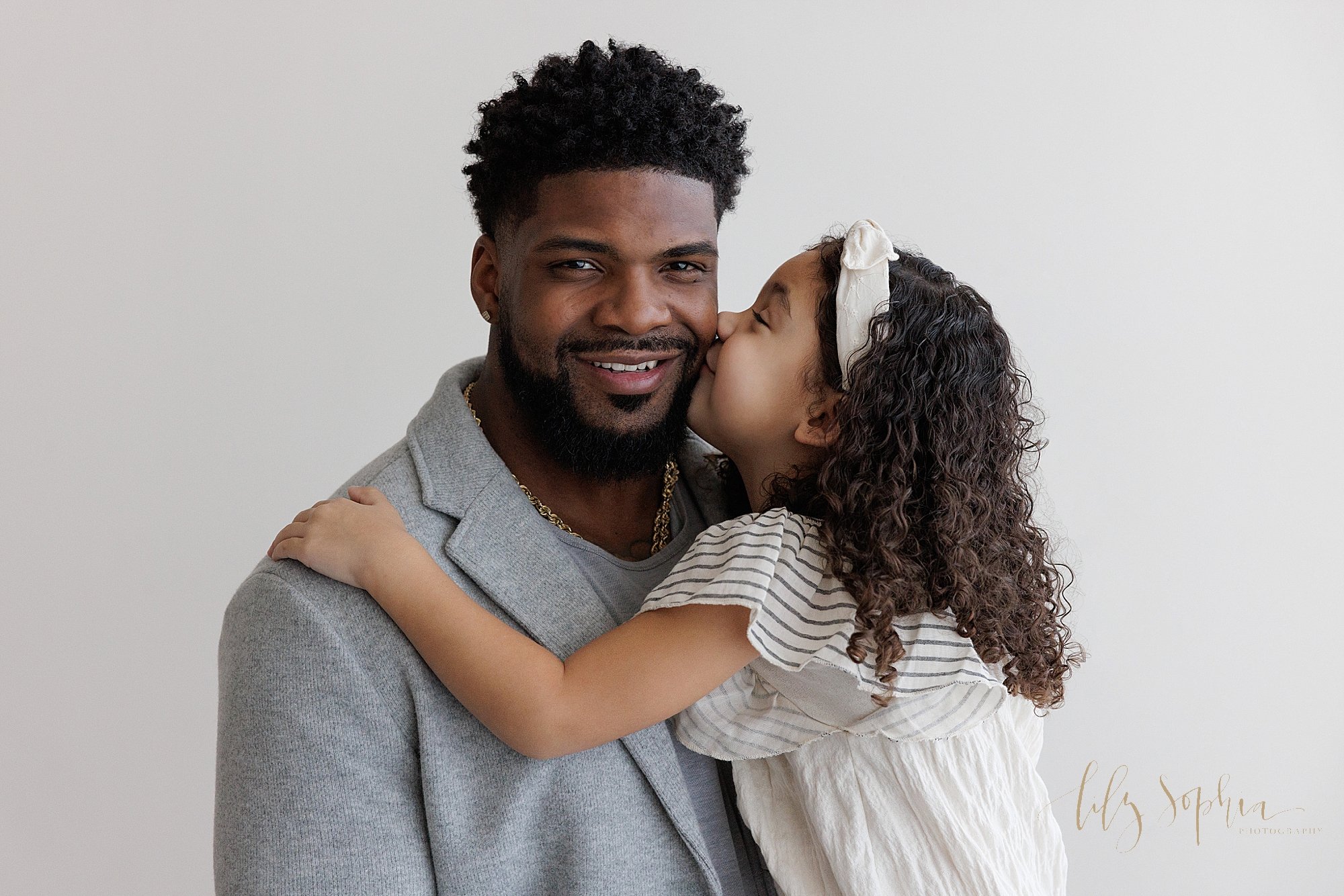  Family photo session with an African-American father as he is kissed on the cheek by his young daughter taken in a photography studio near Poncey Highlands in Atlanta that uses natural light. 