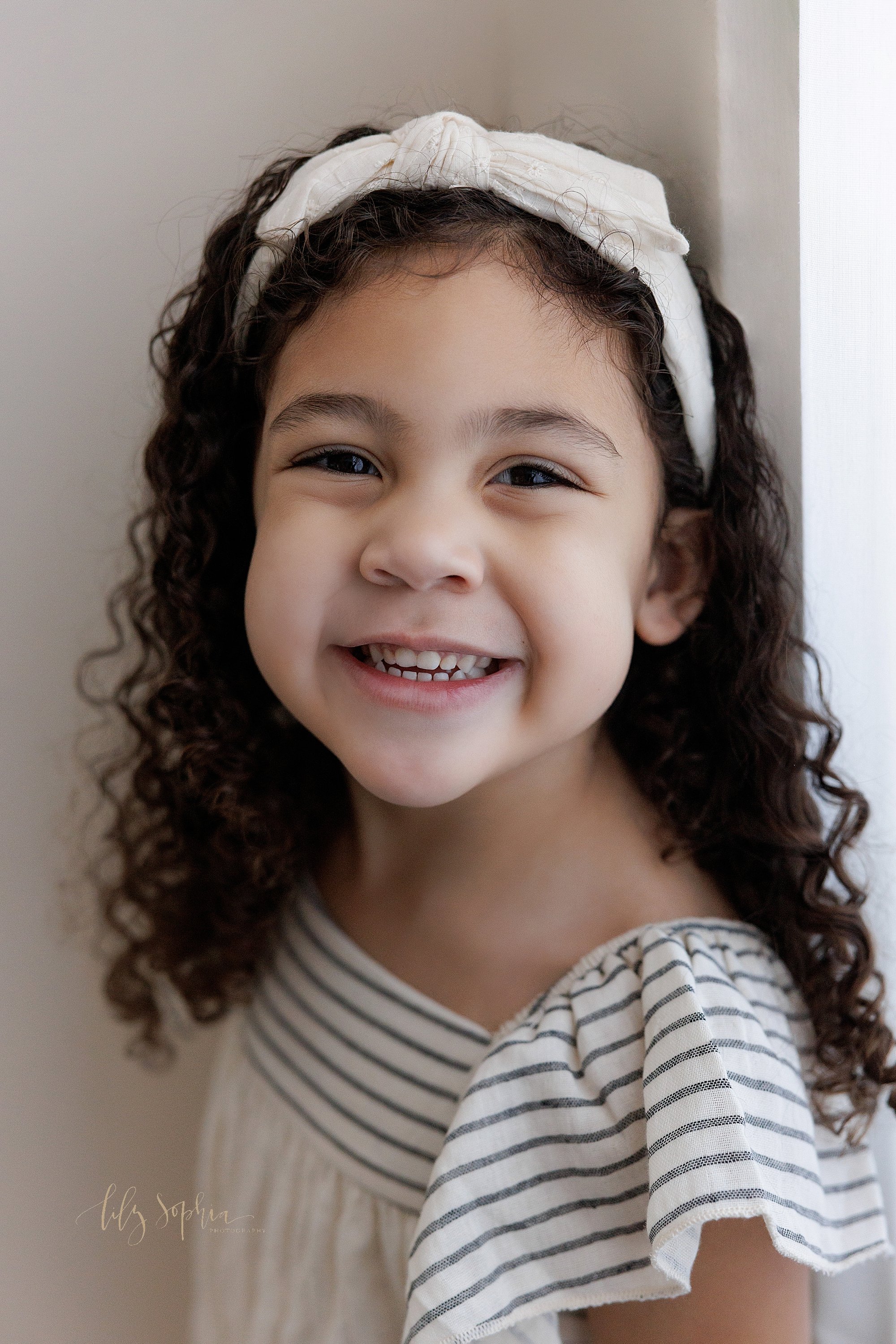  Family portrait of a young girl as she smiles while standing next to a window streaming natural light in a photography studio near Cumming in Atlanta. 