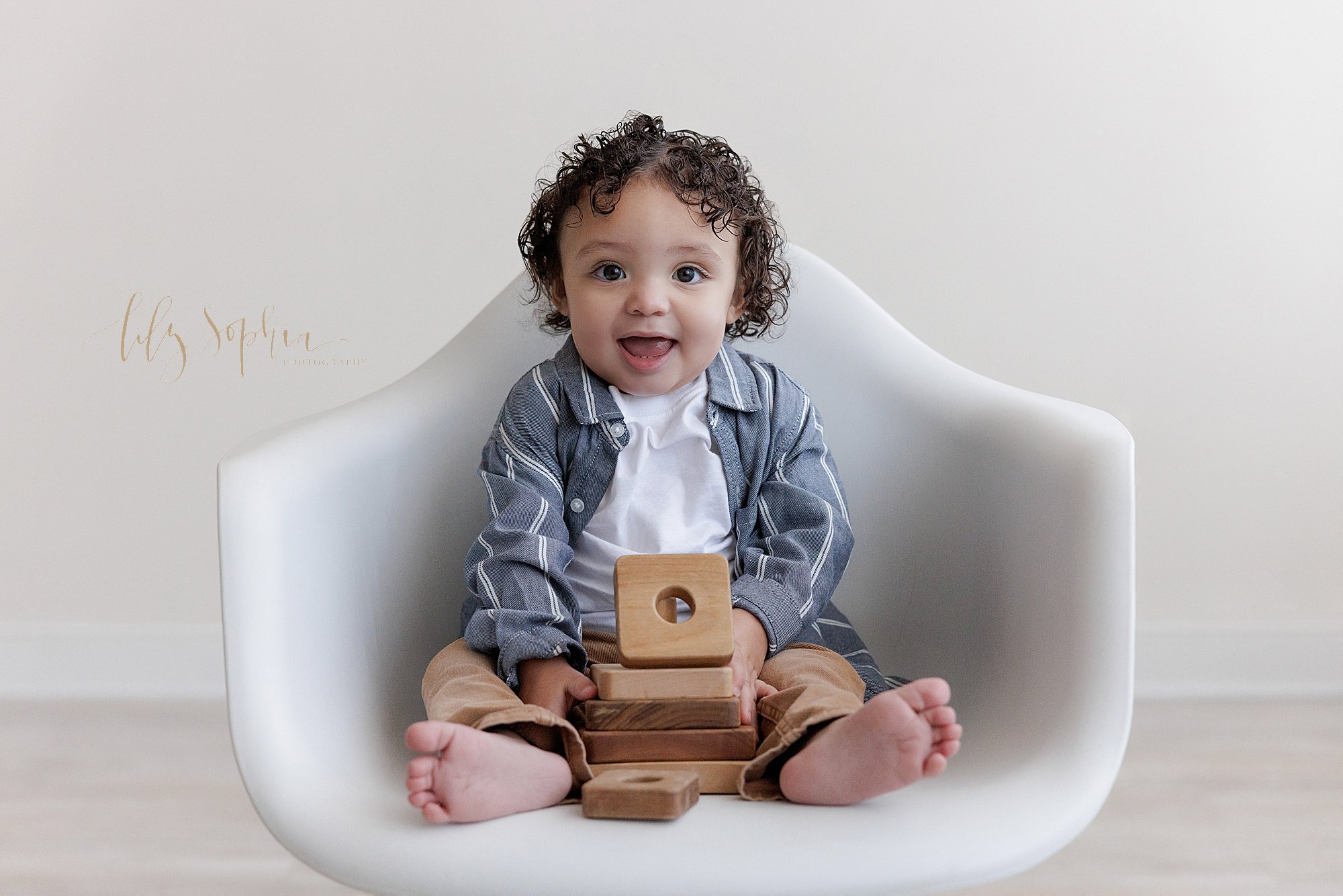  First birthday photo of a one year old little boy as he sits in a white molded chair and plays with a wooden stacking toy taken in a natural light studio near Sandy Springs in Atlanta. 