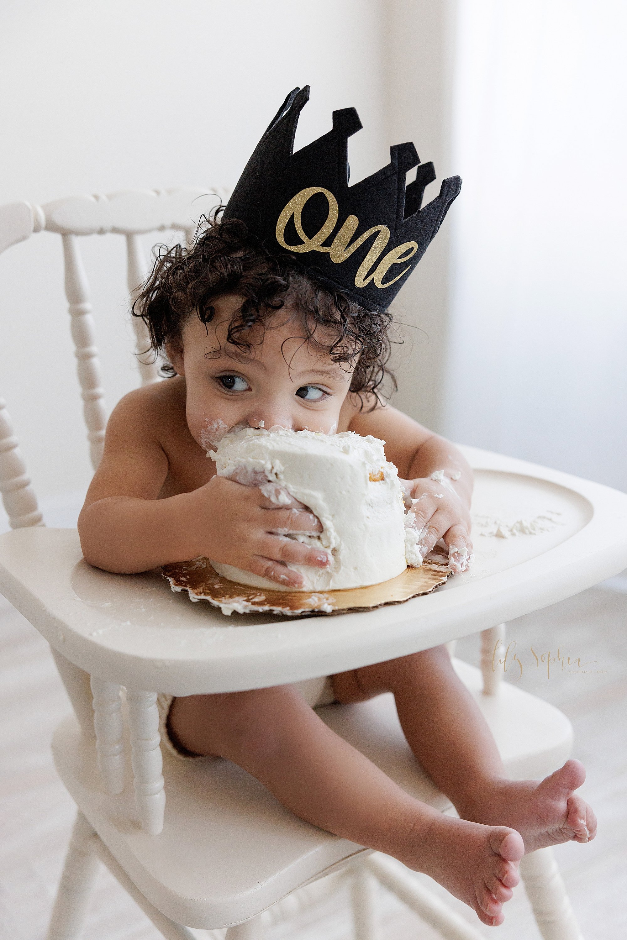  Smash cake first birthday photo session of a one year old little boy wearing a black and gold crown with the number on on it as he holds his cake with his hands and bites into it with his mouth while looking sideways at his parents taken near Buckhe