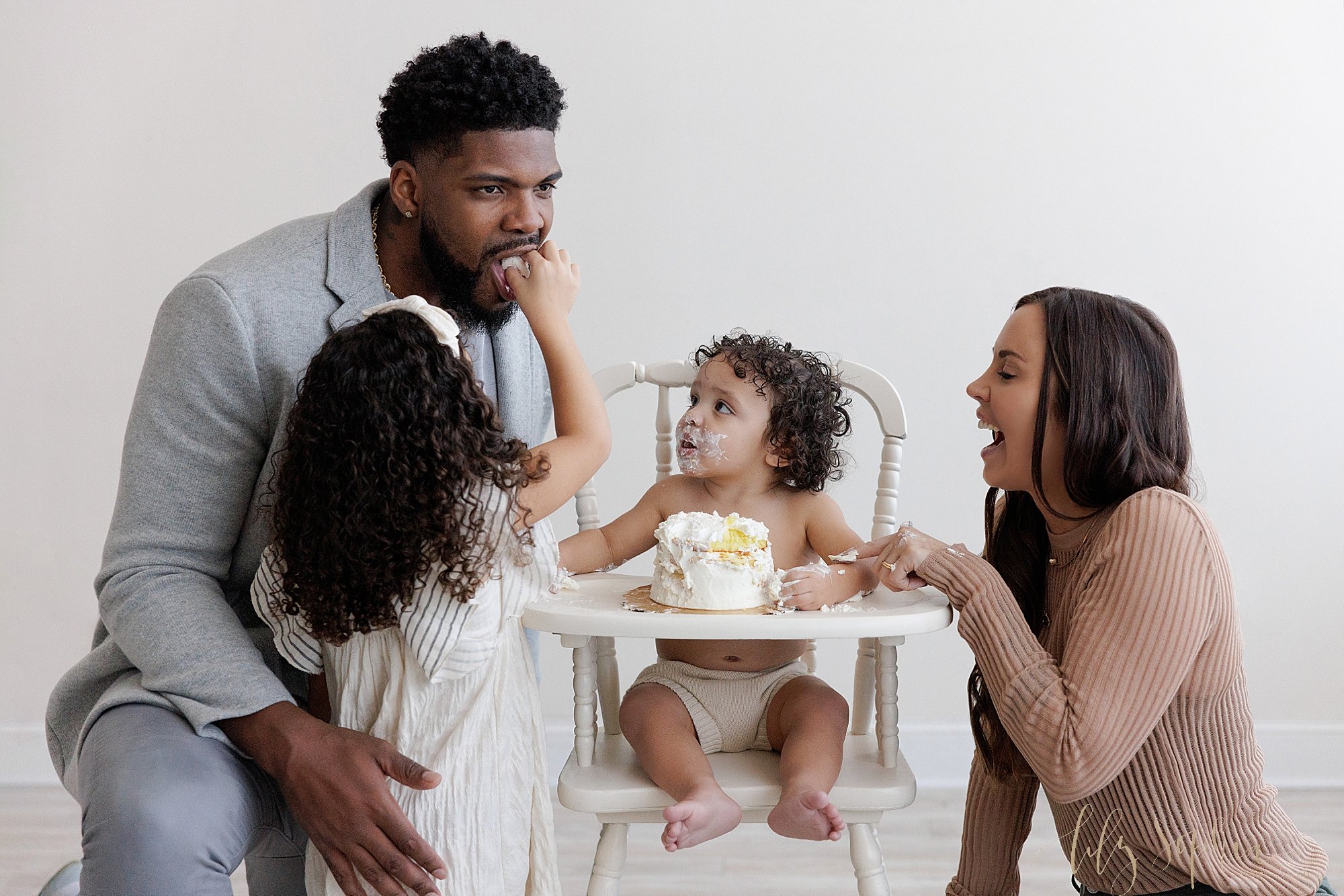  Family first birthday smash cake photo of dad kneeling next to his one year old son who is watching his older sister put a piece of his cake into his dad’s mouth as mom kneels on the other side of the antique high chair the little boy is sitting in 