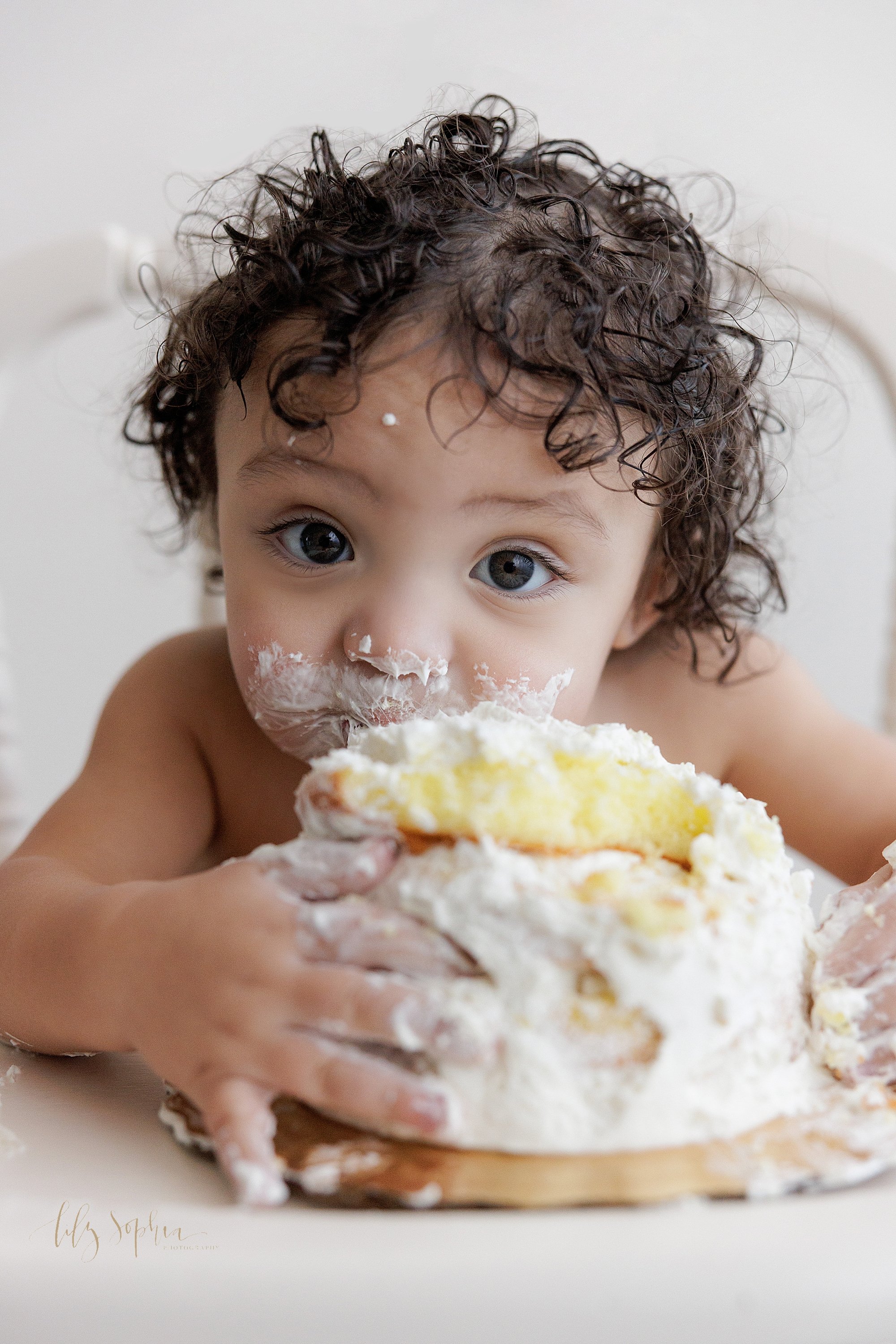  Close-up first birthday portrait of a one year old curly headed boy as he bites into his smash cake with his face covered in icing taken in a photography studio that uses natural light near Ansley Park in Atlanta, Georgia. 