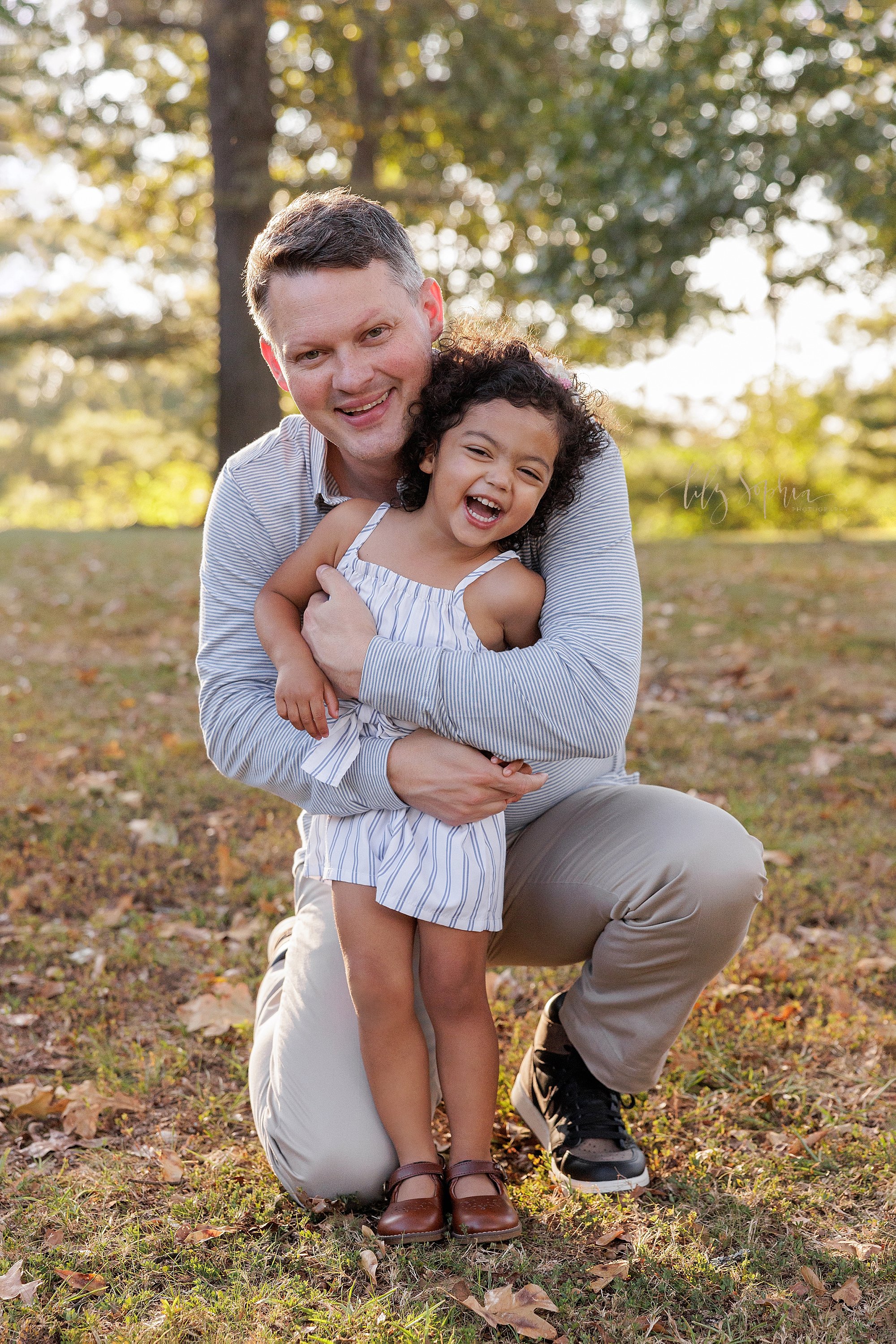  Family picture of a father squatting behind his toddler daughter and hugging her from behind and tickling her as she giggles at sunset in a park near Atlanta during autumn. 