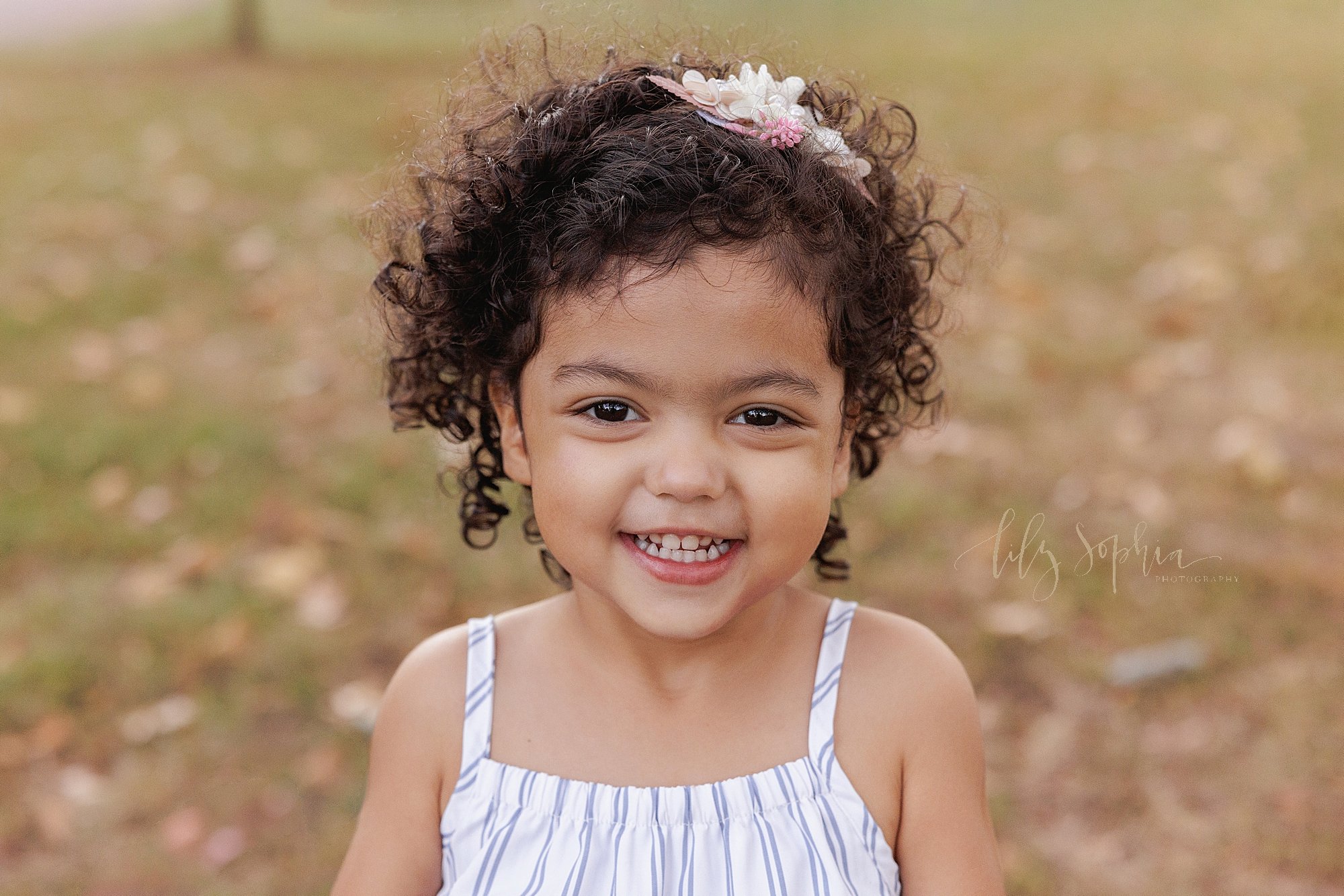  A toddler curly haired toddler girl’s portrait is taken during autumn in Atlanta park as she smiles for a family photo shoot at sunset. 