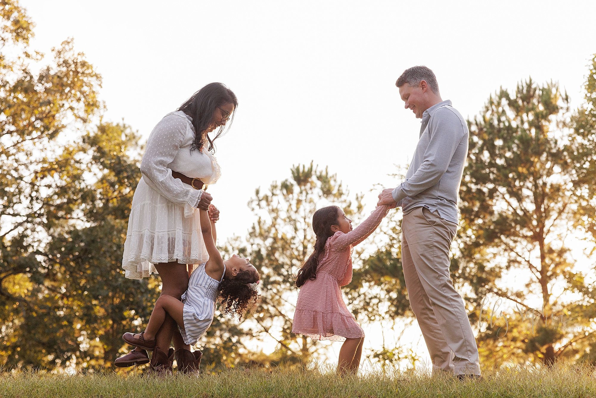  Family photo shoot in a park near Atlanta, Georgia at sunset as dad faces mom and each of the parents play with one their daughters while standing on top of a hill. 