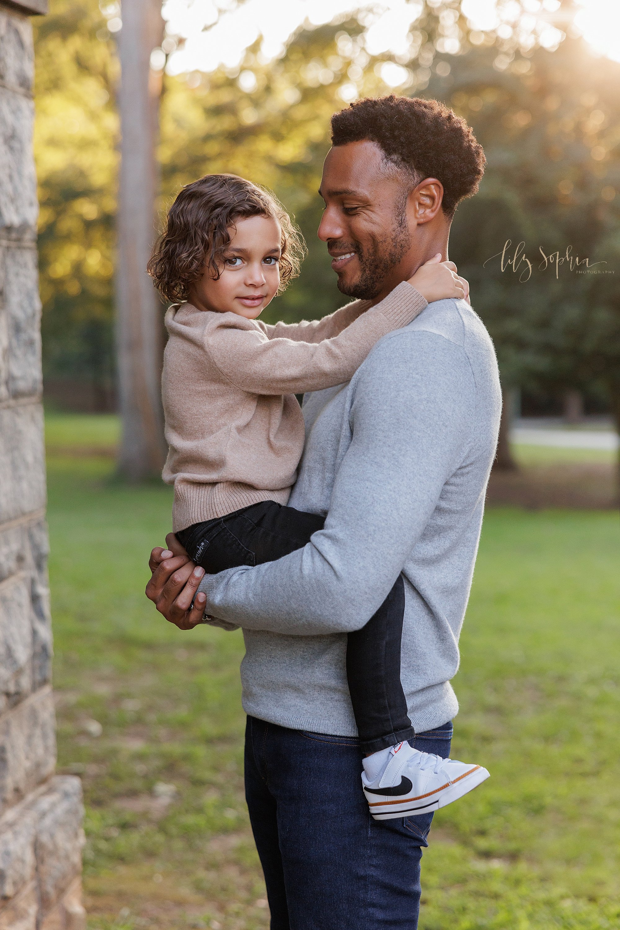  Family portrait of an African-American father as he holds his young son in his arms while he stands at sunset between two stone entranceway pillars in an Atlanta park. 