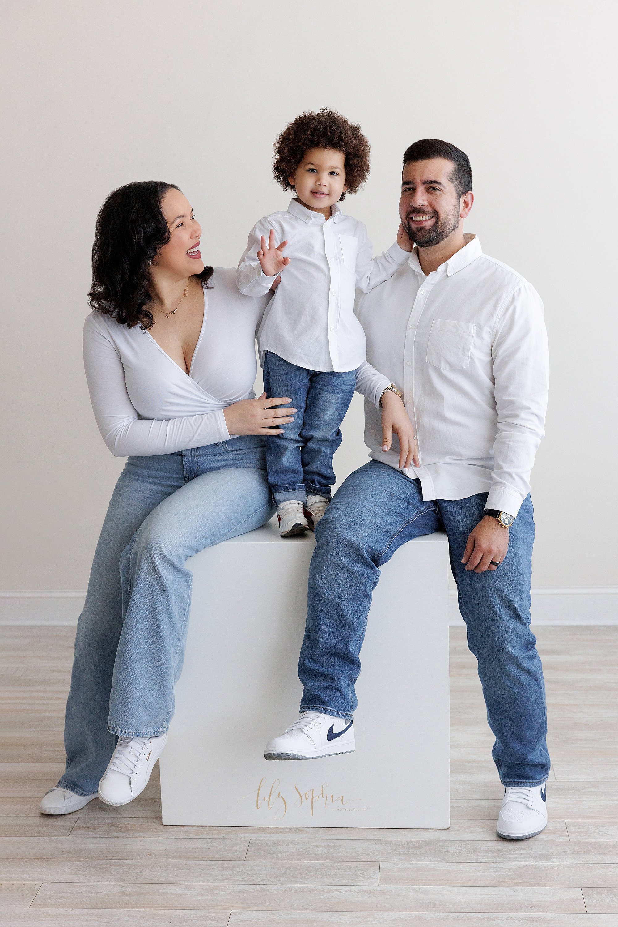  Family photo of a mother and father sitting on a large white cube with their young son standing between them taken using natural light in a photography studio near Oakhurst in Atlanta, Georgia. 