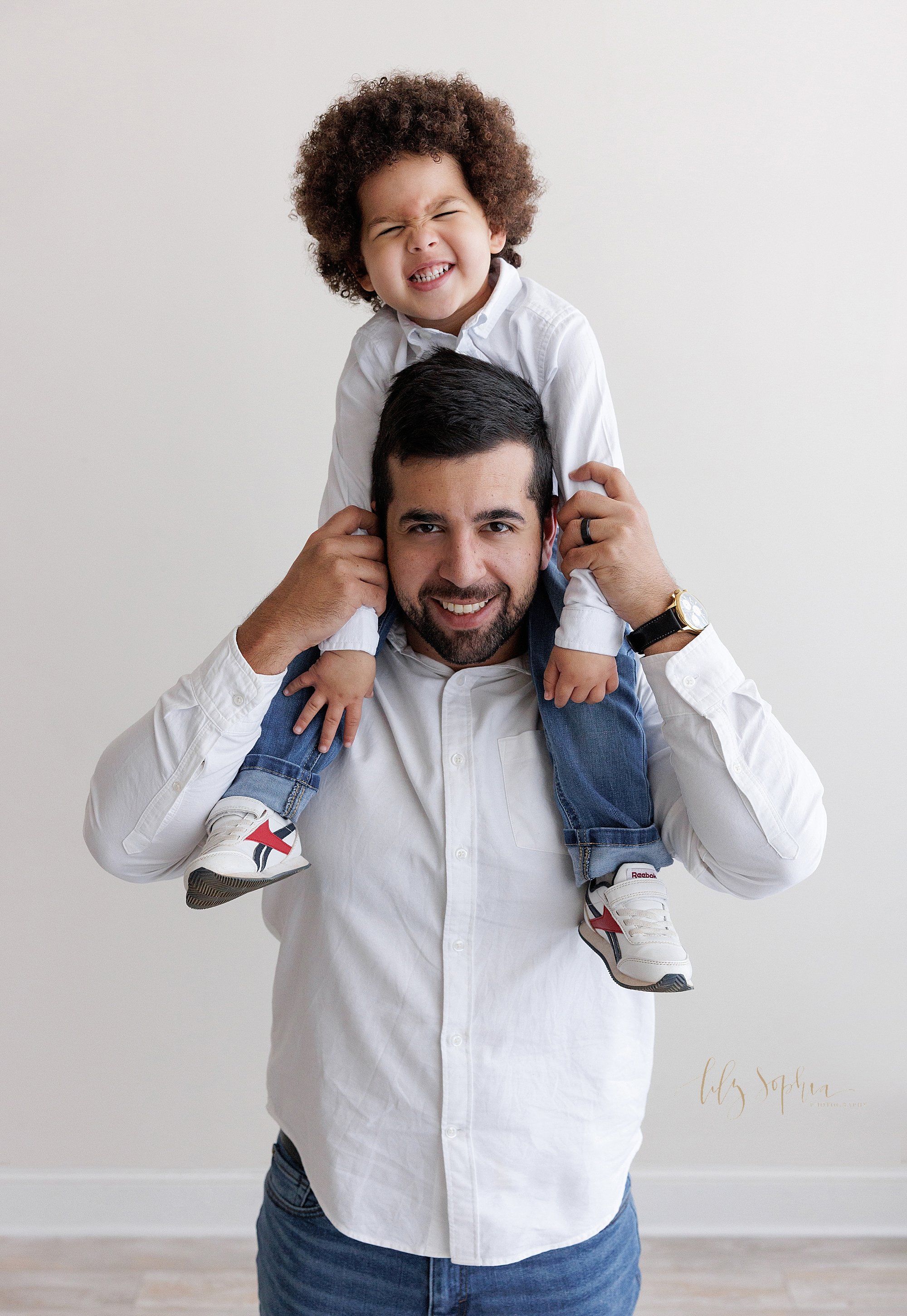  Family photo with a father holding his silly curly haired son on his shoulders as he stands in a photography studio near Morningside in Atlanta in natural light. 