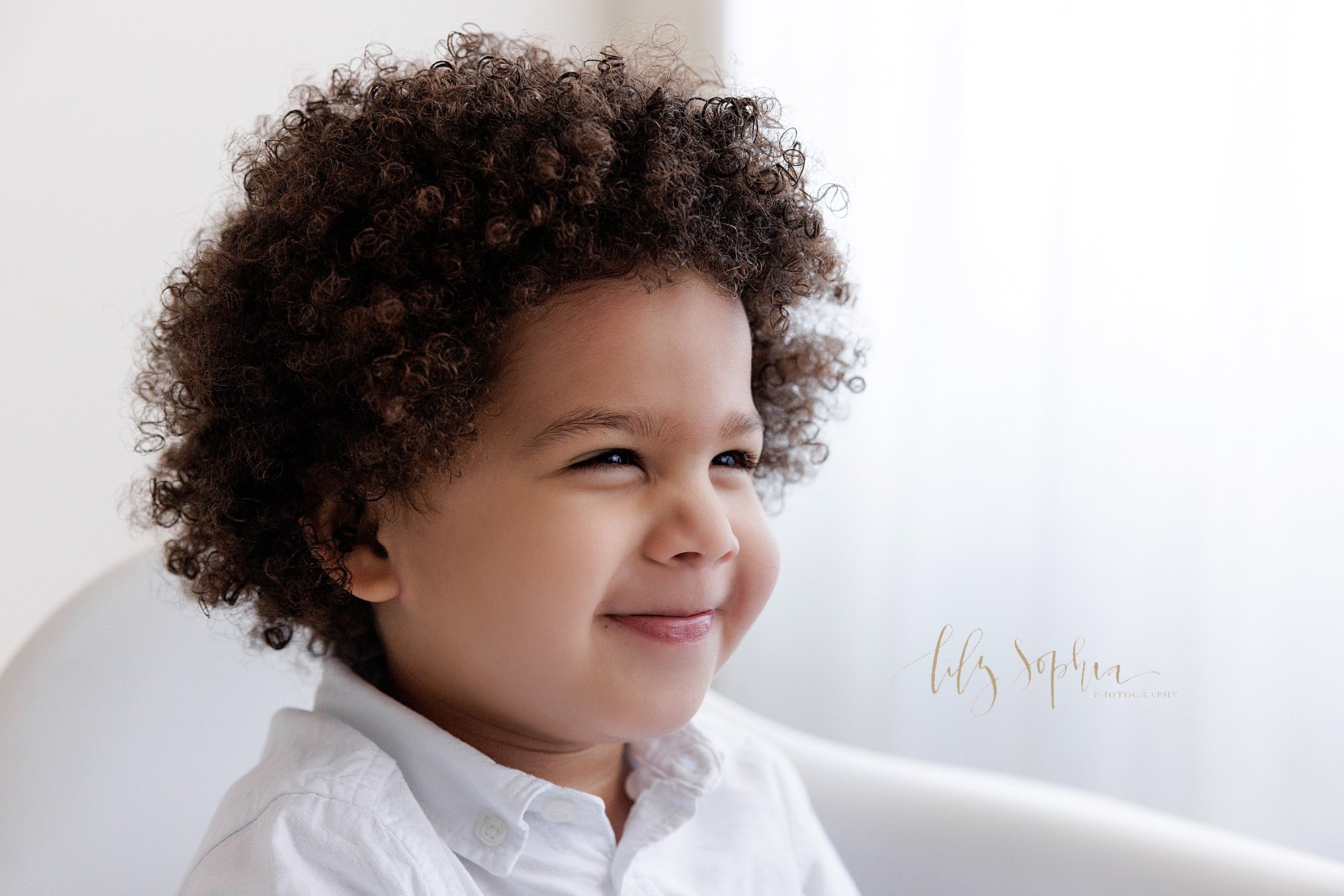  Family photo of a young curly haired boy as he smirks while sitting in a white molded chair next to a window streaming natural light in a photography studio near Buckhead in Atlanta. 