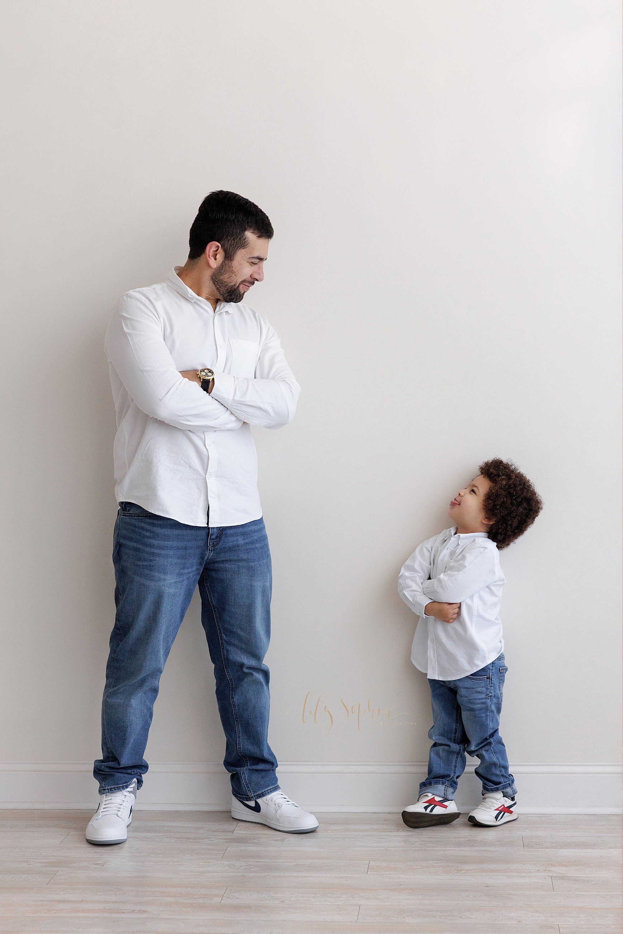  Family father and son picture with the father standing with his arms folded across his chest as he turns his head over his left shoulder to look down at his young son who is standing and folding his arms over his chest and looks up to his father tak