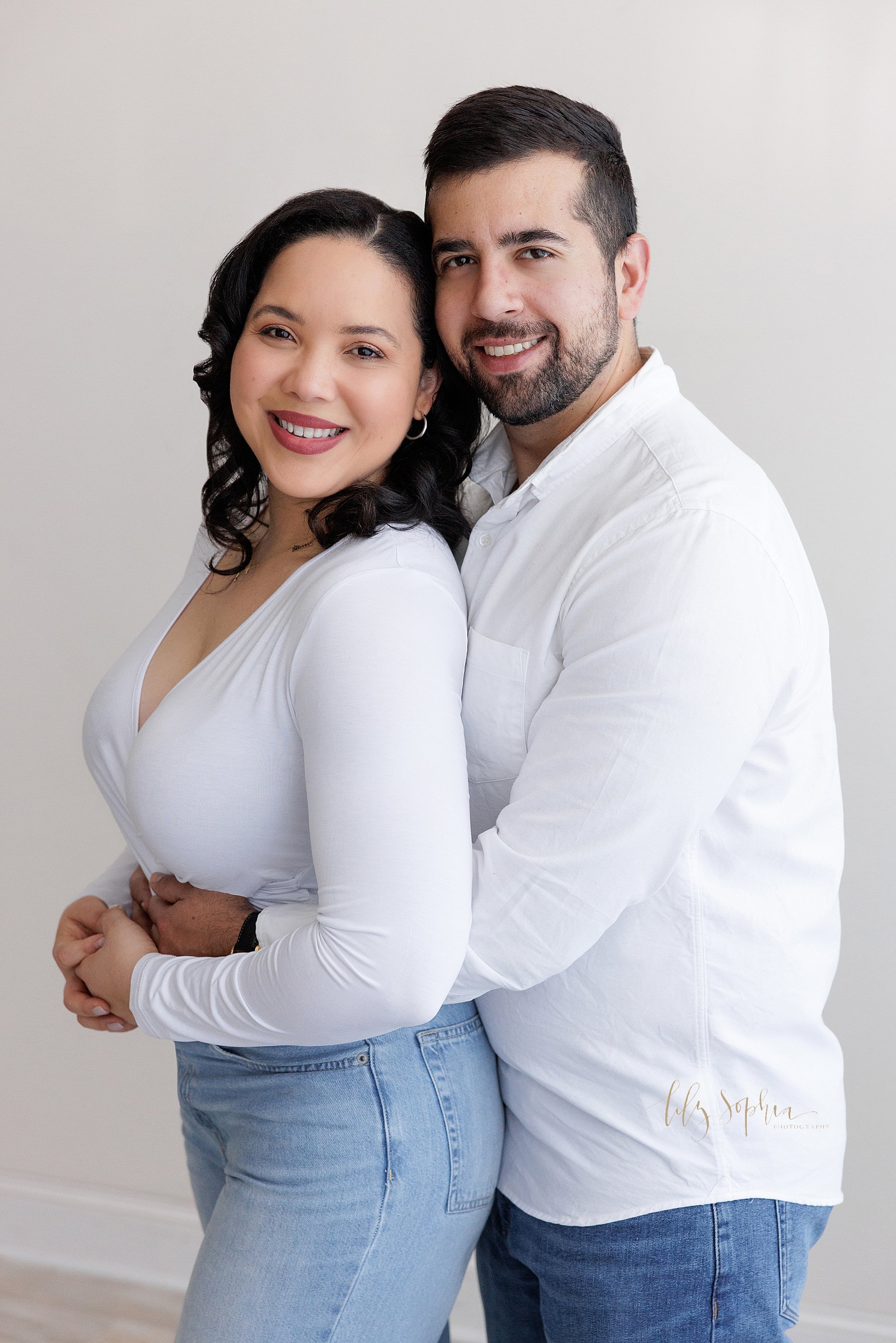  Family photo of a husband and wife as they stand in a photography studio with the husband’s chest against his wife’s back as he hugs her around the waist in natural light ner Alpharetta in Atlanta, Georgia. 