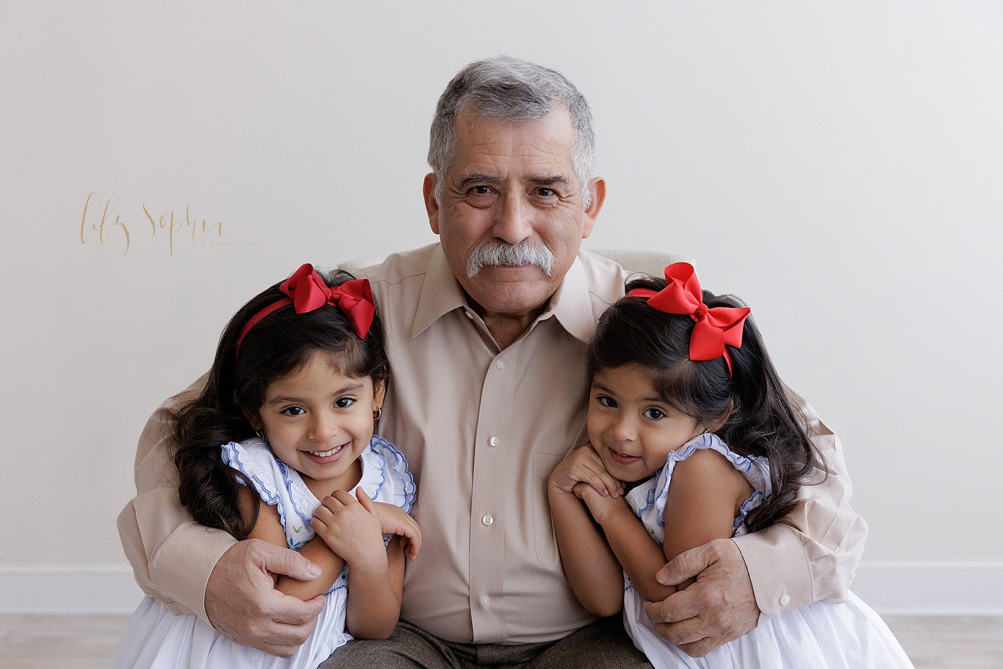  Family portrait of a grandfather sitting on a tufted bench in a natural light studio with his granddaughters on either side of him as he squeezes them tight taken near Virginia Highlands in Atlanta, Georgia. 