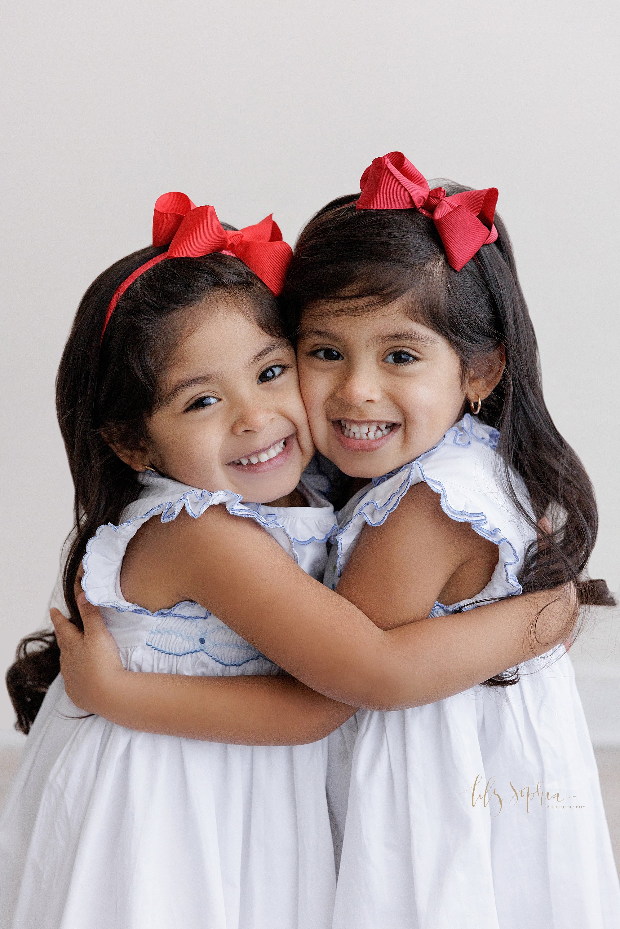  Family photo of sisters as they hug one another with their faces cheek to cheek taken in natural light near Smyrna in Atlanta in a photography studio. 