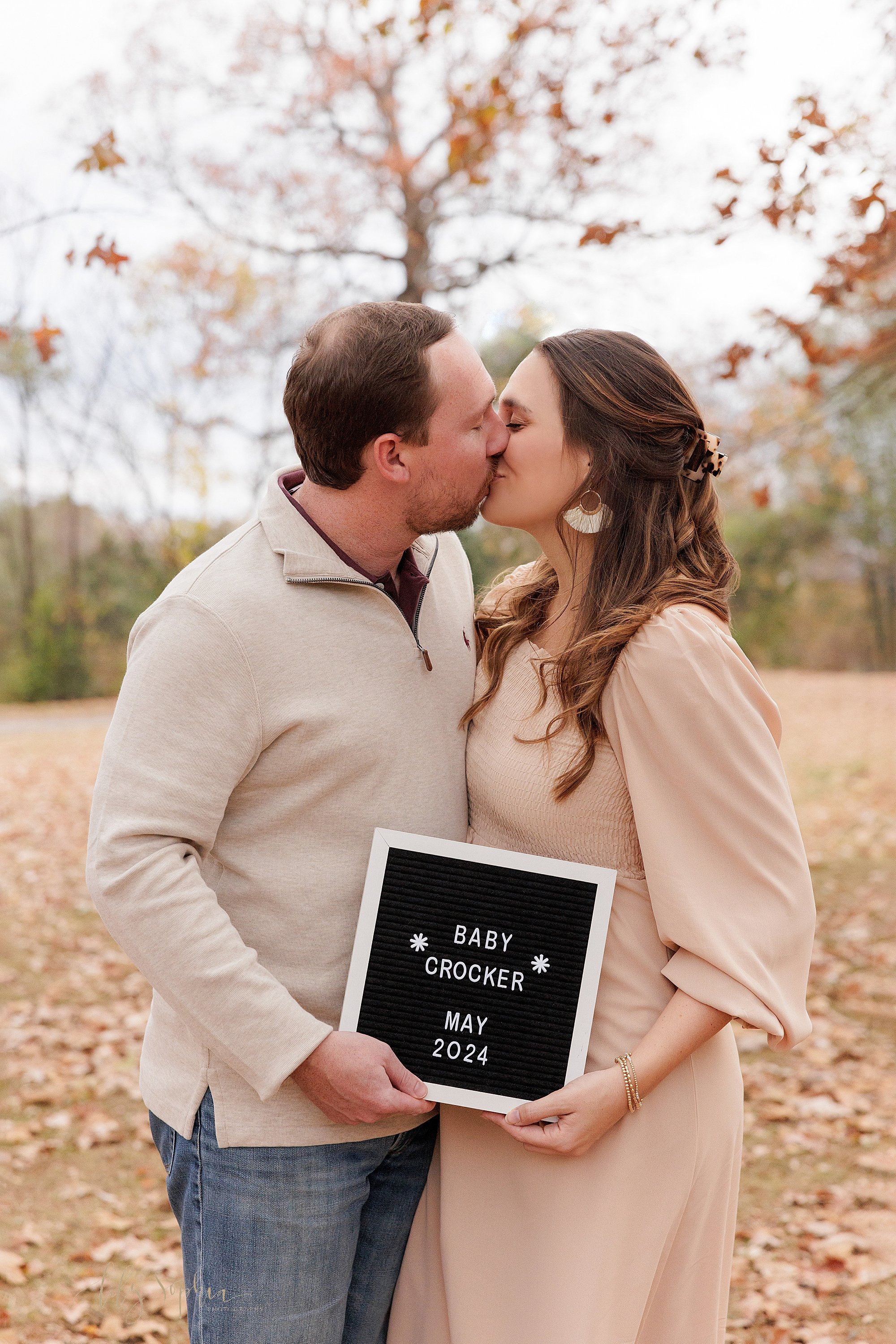  Maternity couples of a mother and father as they kiss one another while standing in an Atlanta park during autumn at sunset holding their announcement board to announce their pregnancy. 