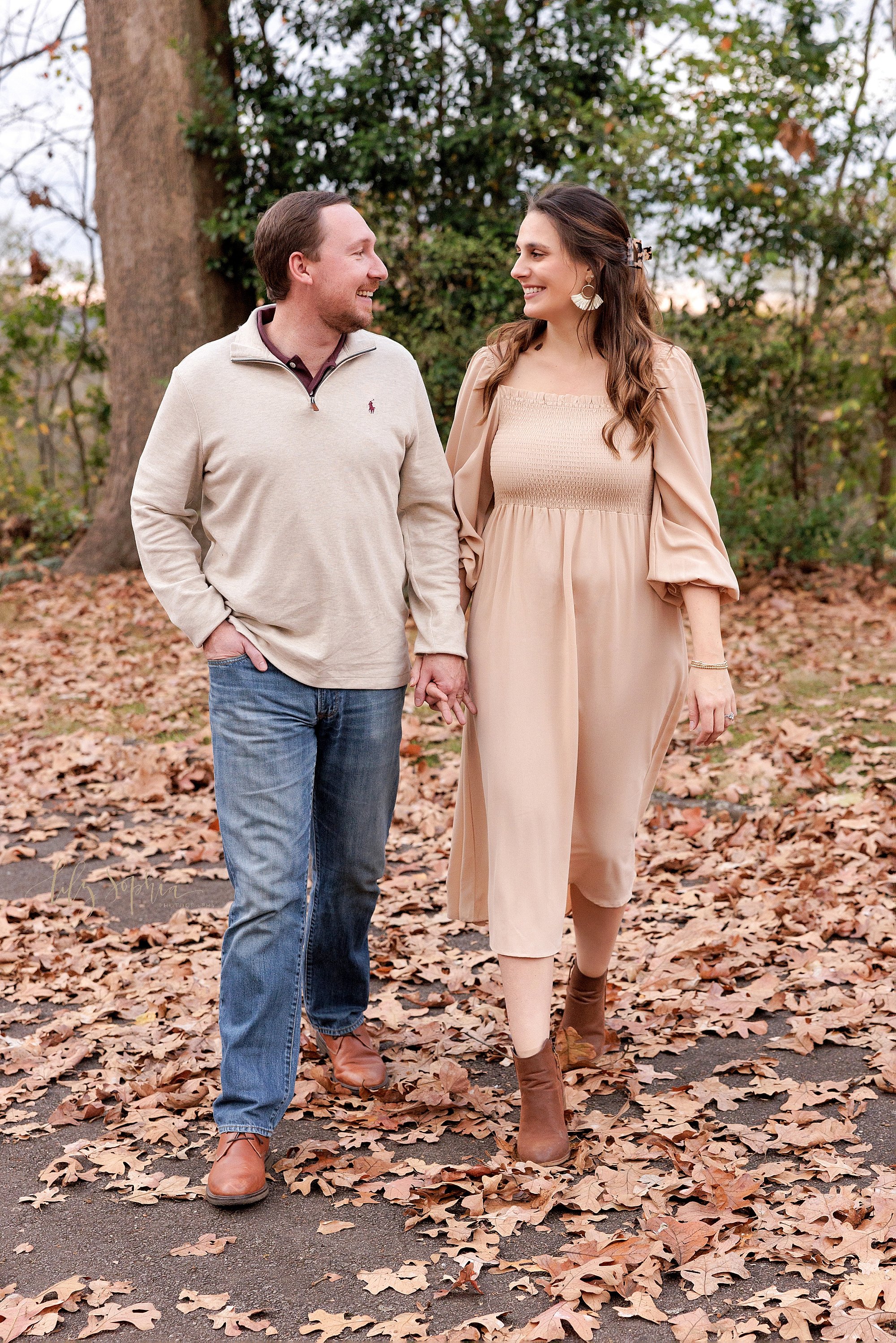  Maternity photo shoot in an Atlanta park at sunset while the couple talk to one another while walking hand in hand along a leaf covered path during autumn. 