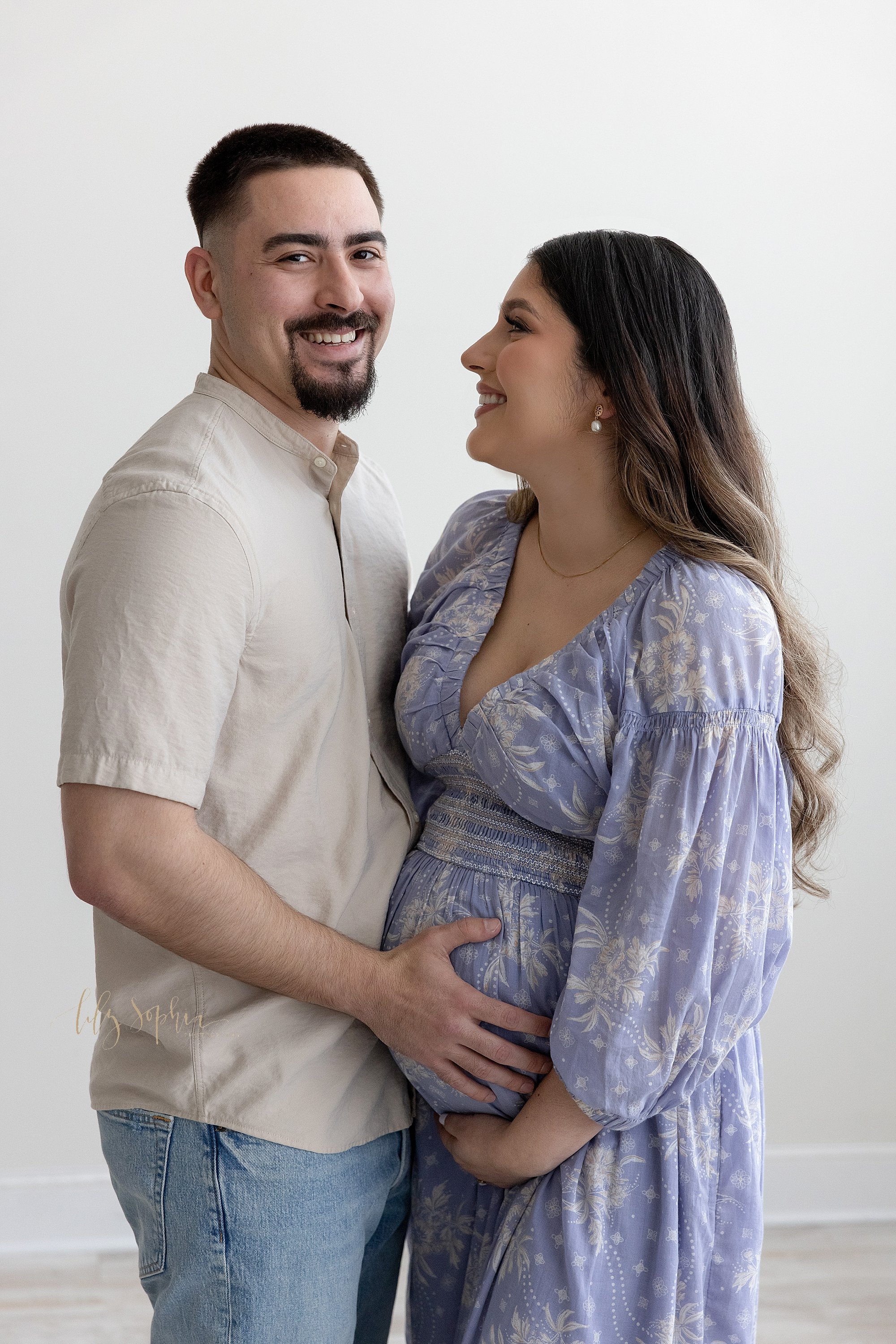  Maternity portrait of a mother and father as they face one another with mom holding the base of her belly and dad placing his right hand on her belly as the father turns his head to the right and his wife smiles at him taken in a natural light studi